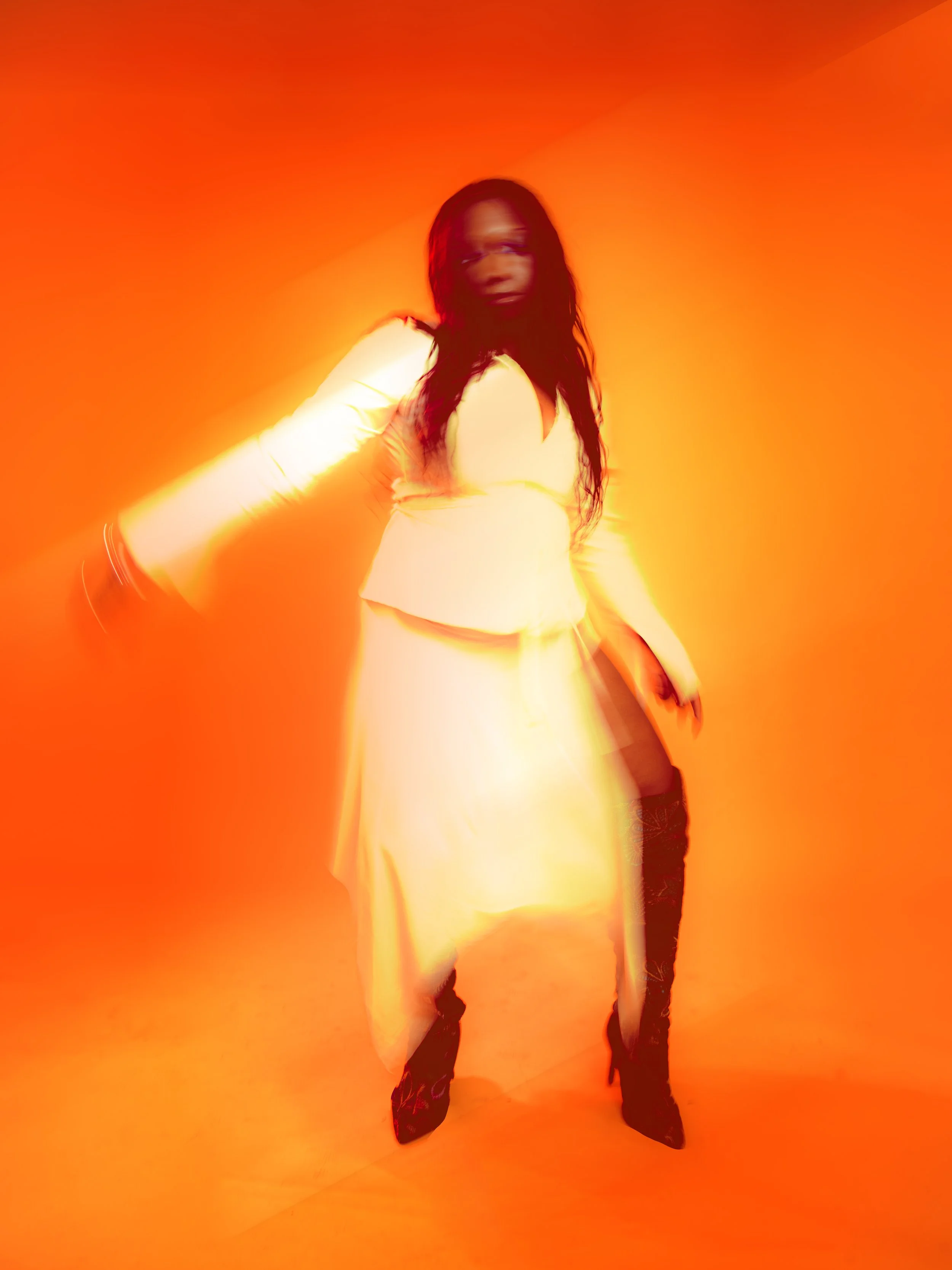 Local Dallas, Texas, DFW Non-traditional Editorial Creative Portraits Cover Art Photographer. Musician Courtney Noir. A woman in a light-colored dress and black high-heeled boots stands against a vibrant orange background, with her face slightly blur