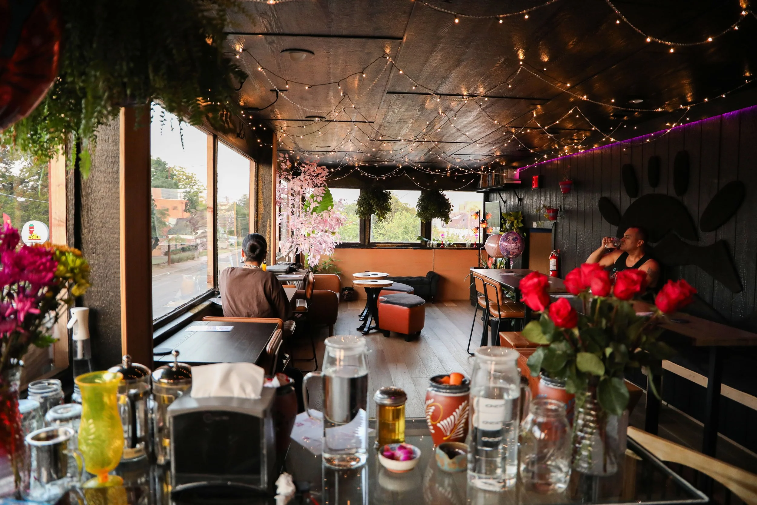 Interior of a cozy restaurant with wooden walls decorated with string lights. Large windows reveal a street view outside. There are tables and chairs, with some patrons sitting and drinking. The foreground features a table with various items includin