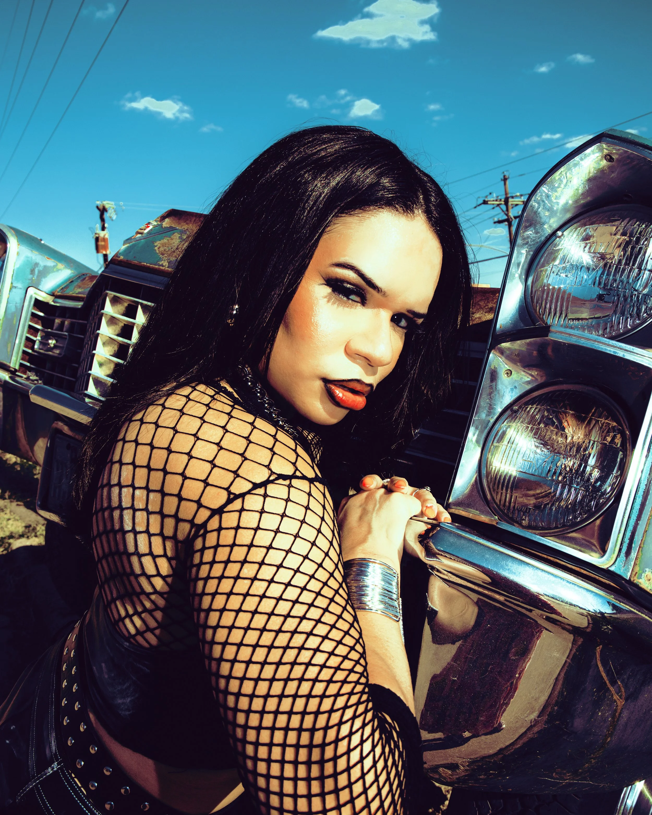 Local Dallas, Texas, DFW Non-traditional Fashion Editorial Creative Portraits Cover Art Photographer Photoshoot. @roseypears . A woman with dark hair, wearing makeup and fishnet clothing, poses near a chrome vehicle under a blue sky with a few clouds