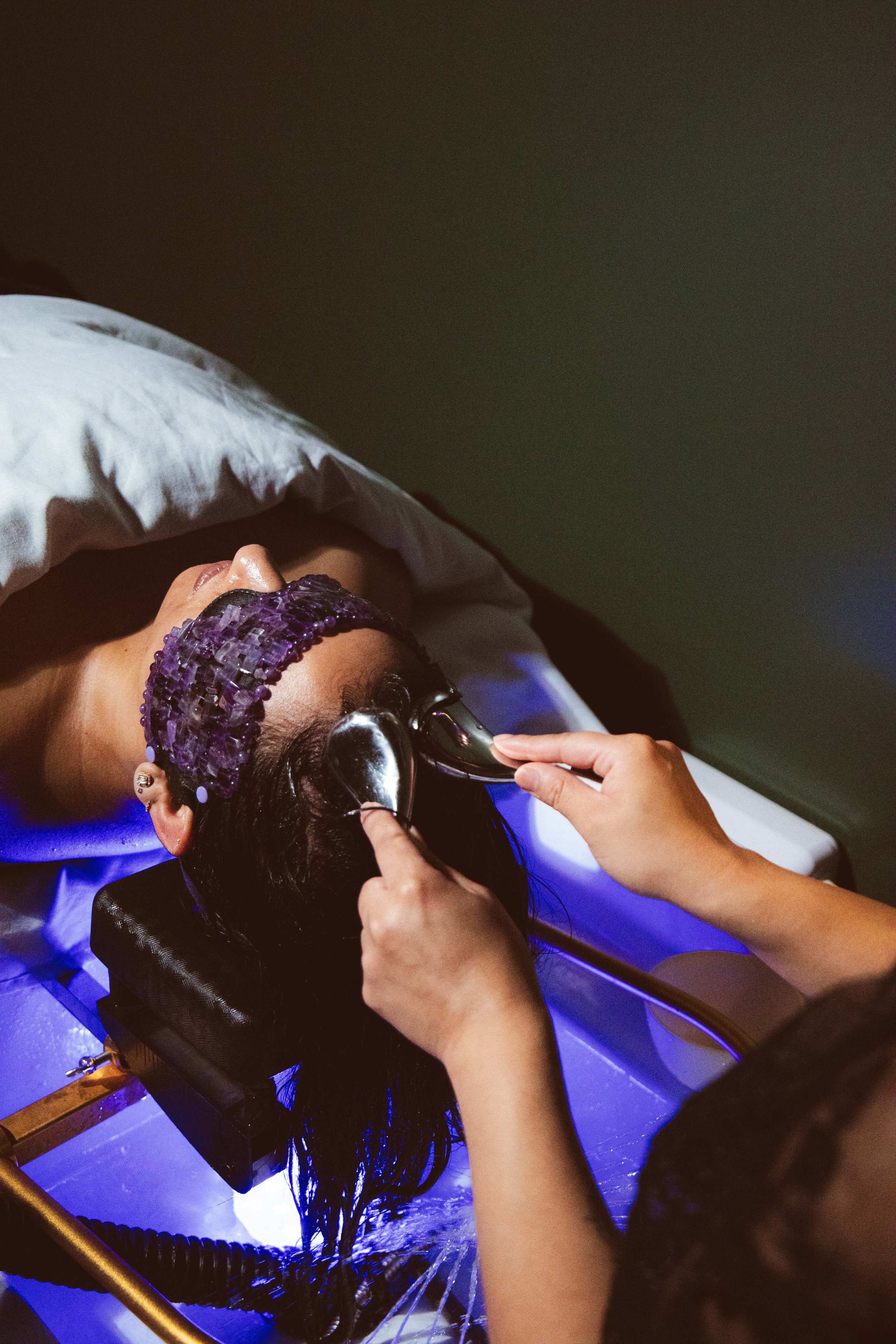 A person lying on a reclined chair receiving a facial treatment, wearing a purple headband, with a white towel covering their body.