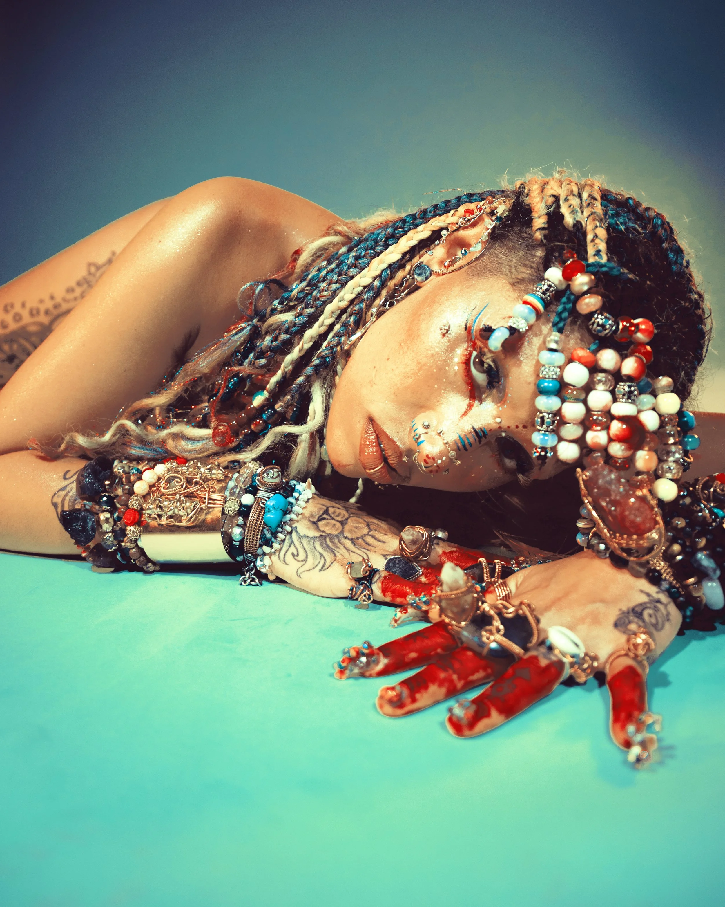 Local Dallas, Texas, DFW Non-traditional Fashion Editorial Creative Portraits Cover Art Photographer Photoshoot. A person lying on a turquoise surface, adorned with colorful jewelry and intricate face and body paint, with a gradient background.