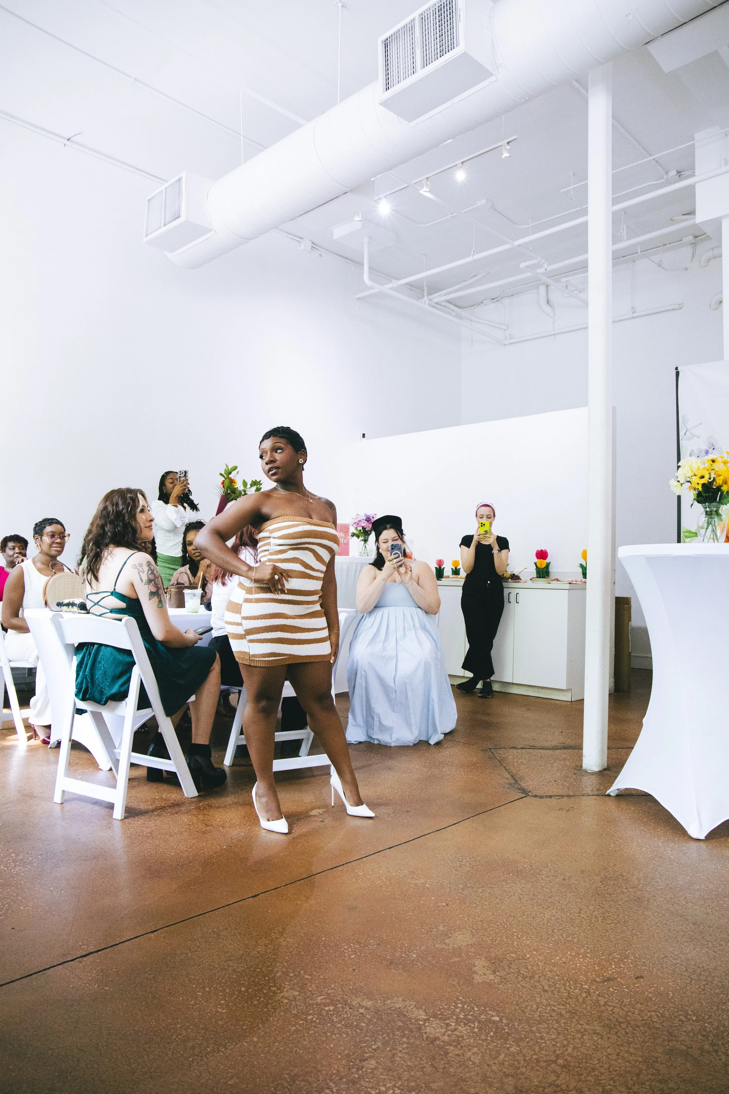 A woman in high heels and a strapless zebra print dress standing confidently in a brightly lit room with seated and standing guests, some taking photos, decorated with colorful flowers on tables.