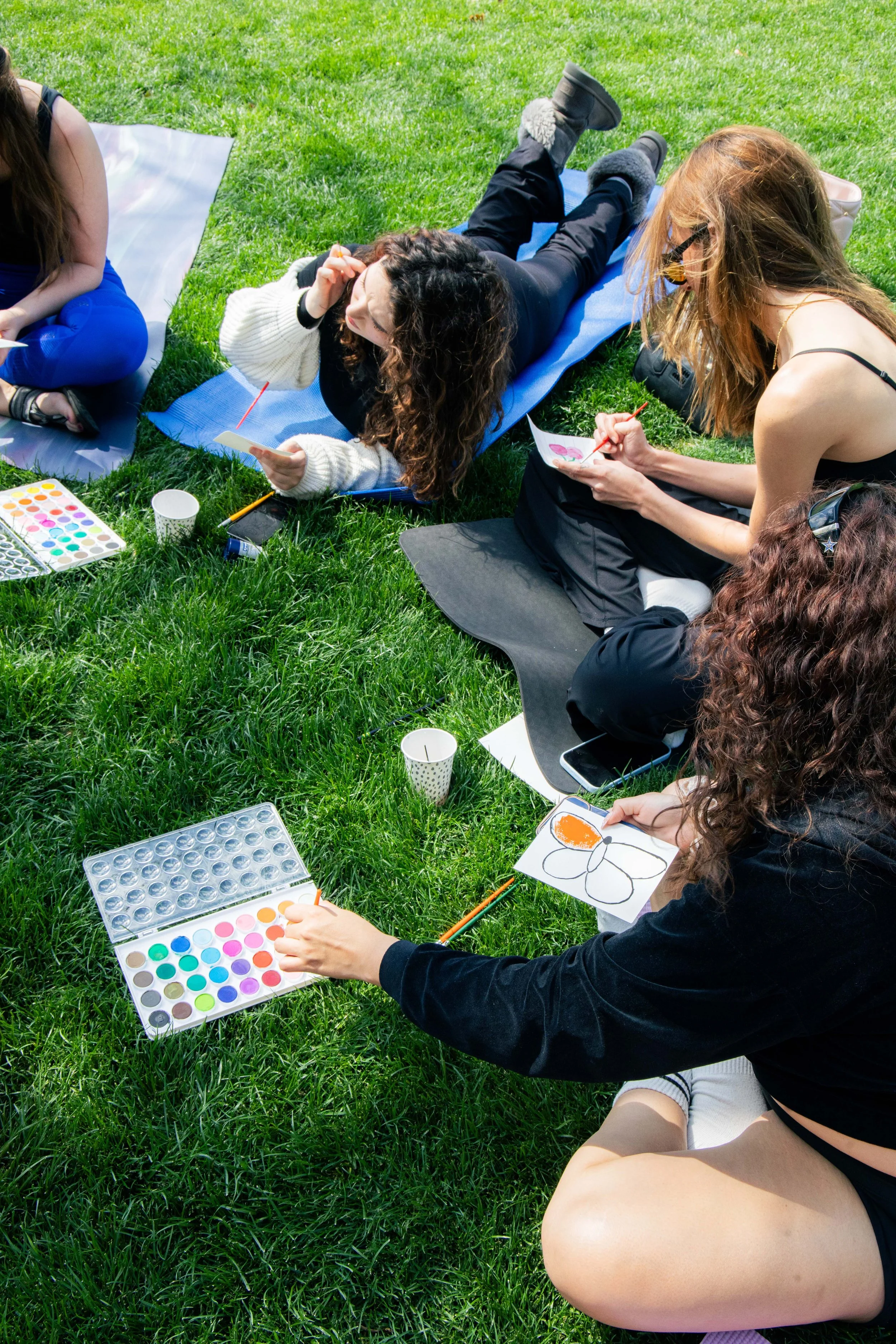 Group of women painting and drawing on paper while sitting on the grass outdoors on a sunny day.