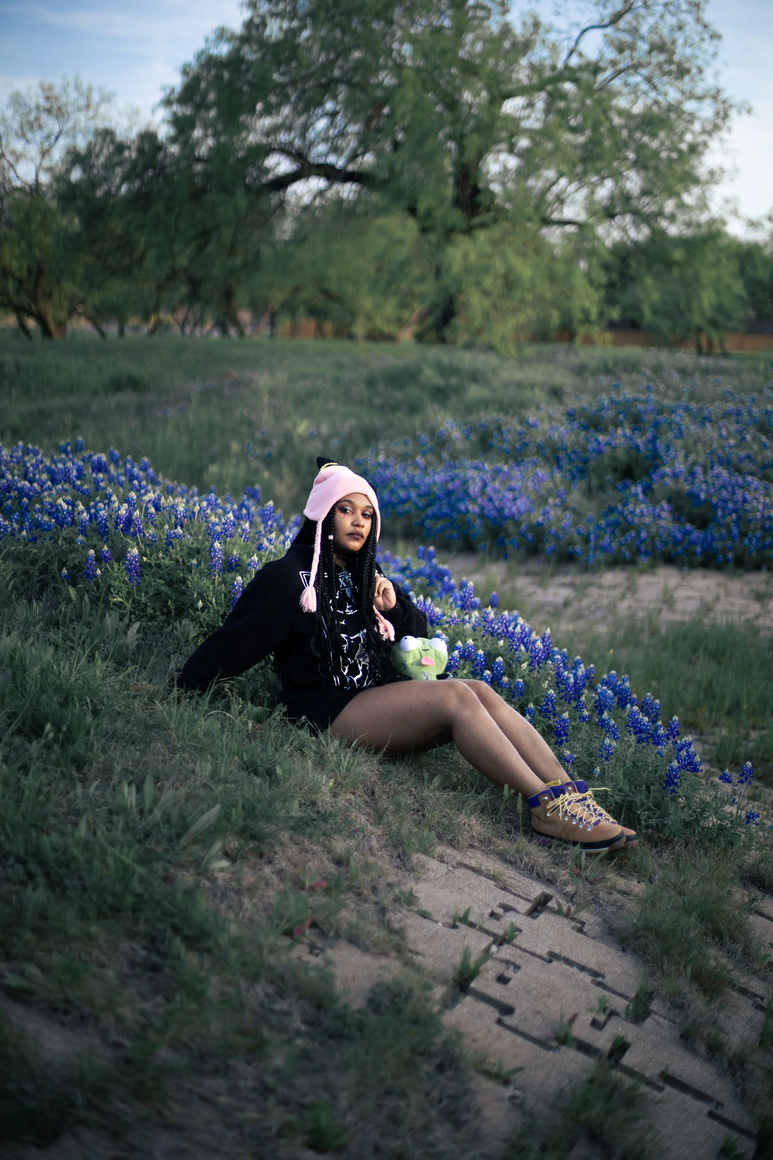 Local Dallas, Texas, DFW Non-traditional Fashion Editorial Creative Portraits Cover Art Photographer Photoshoot. Musician Igimeji . A young woman with long braided hair sitting on the grass near blue flowers in a park, wearing a pink beanie hat, blac