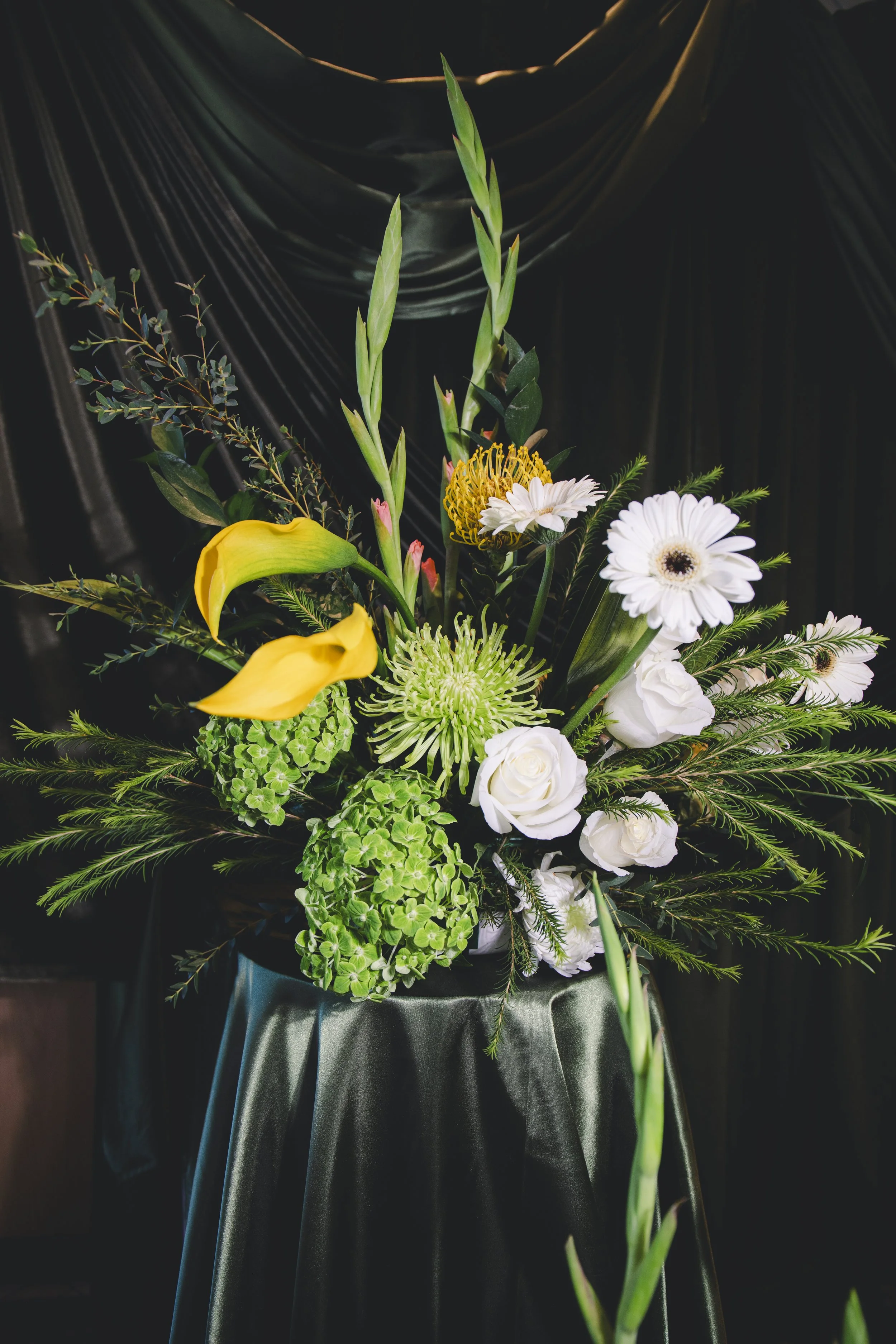 A bouquet of various flowers including white daisies, yellow calla lilies, and green foliage against a black background.