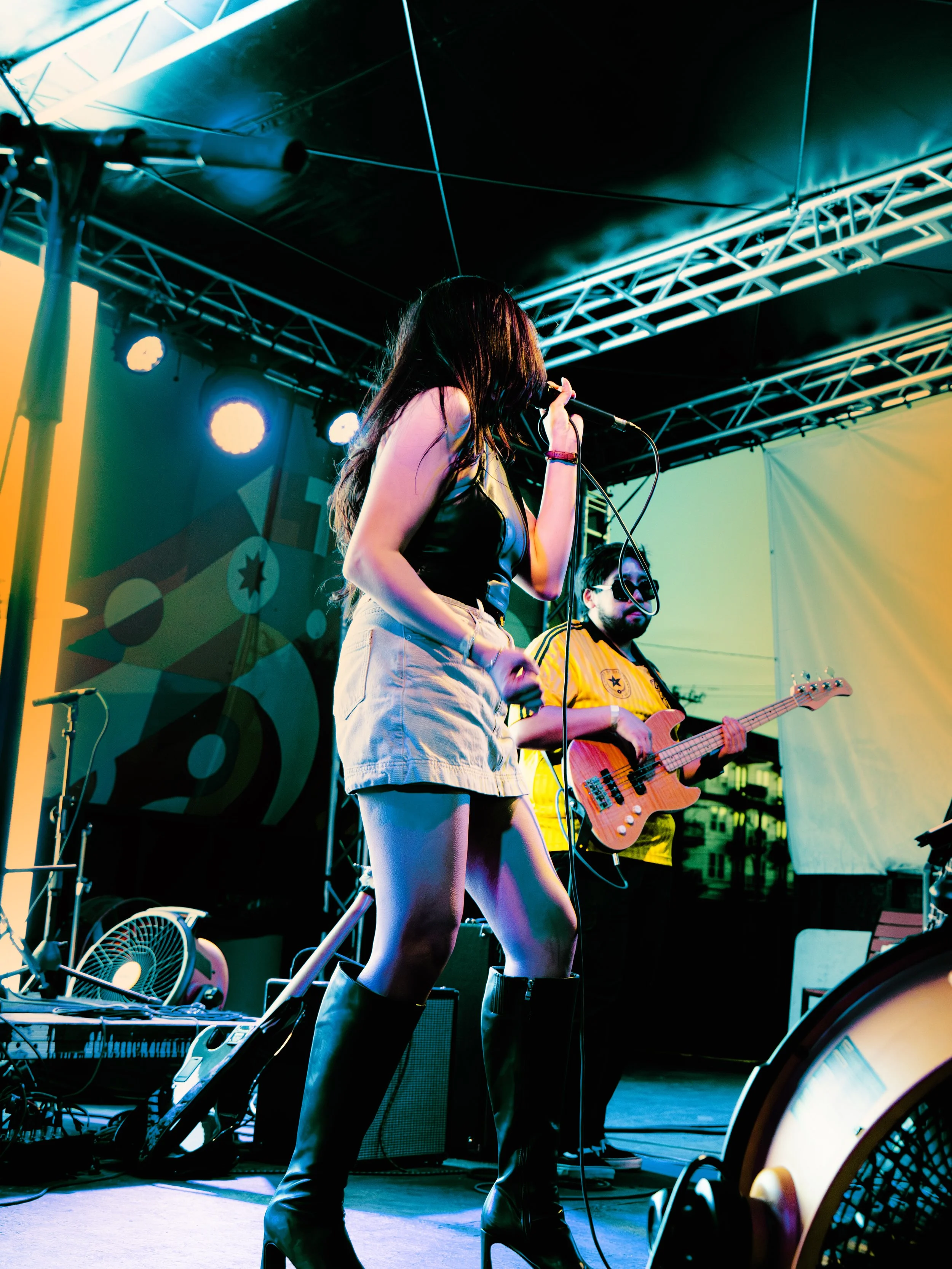 A female singer with long hair wearing black boots and a sleeveless top performs on stage, accompanied by a male bassist in a yellow shirt and sunglasses. The stage has colorful lights and a backdrop with circular patterns.