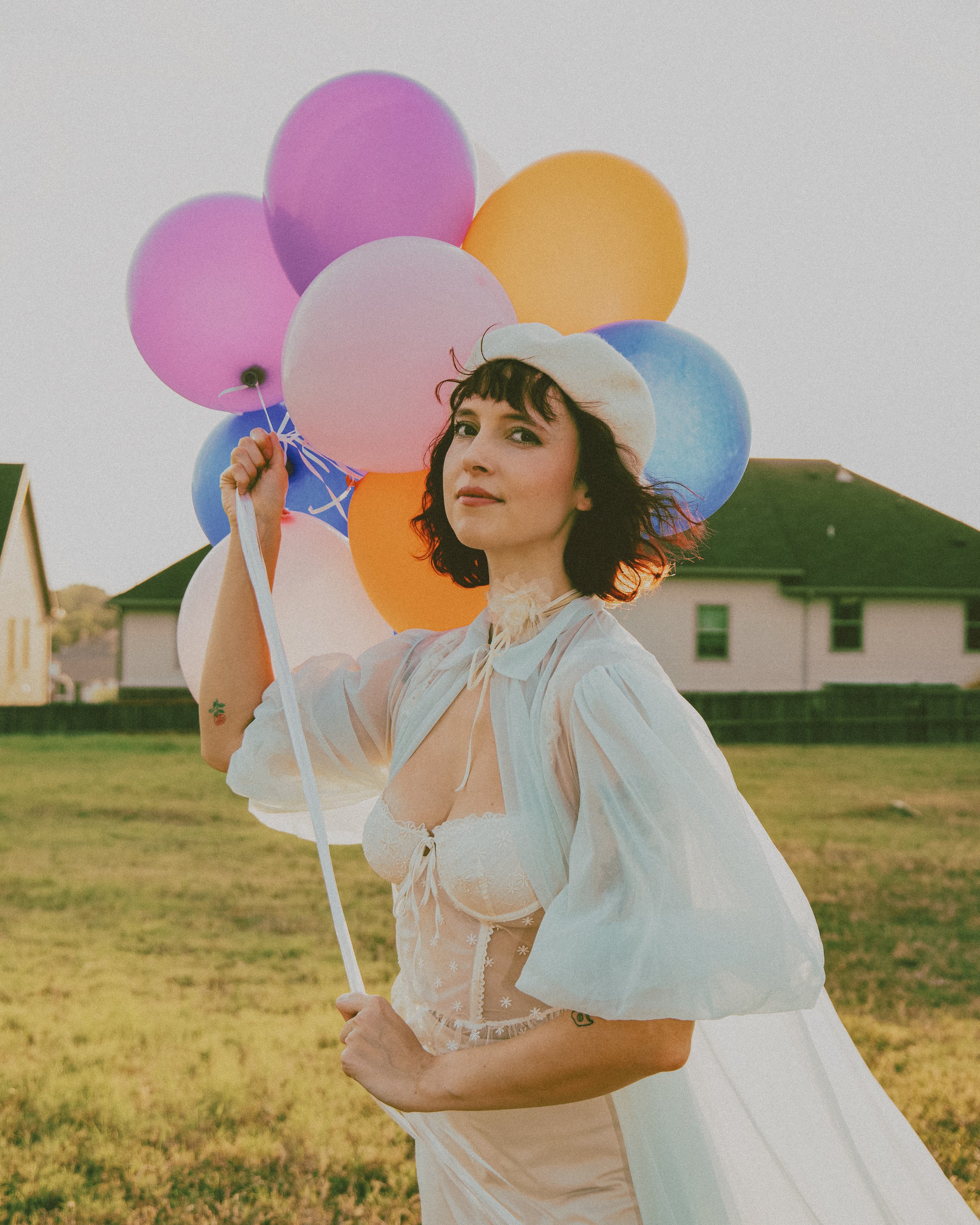 Local Dallas, Texas, DFW Non-traditional Fashion Editorial Creative Portraits Cover Art Photographer Photoshoot. A woman with dark, curly hair holding colorful balloons outdoors, wearing a white beret and a sheer, white, puffy blouse.