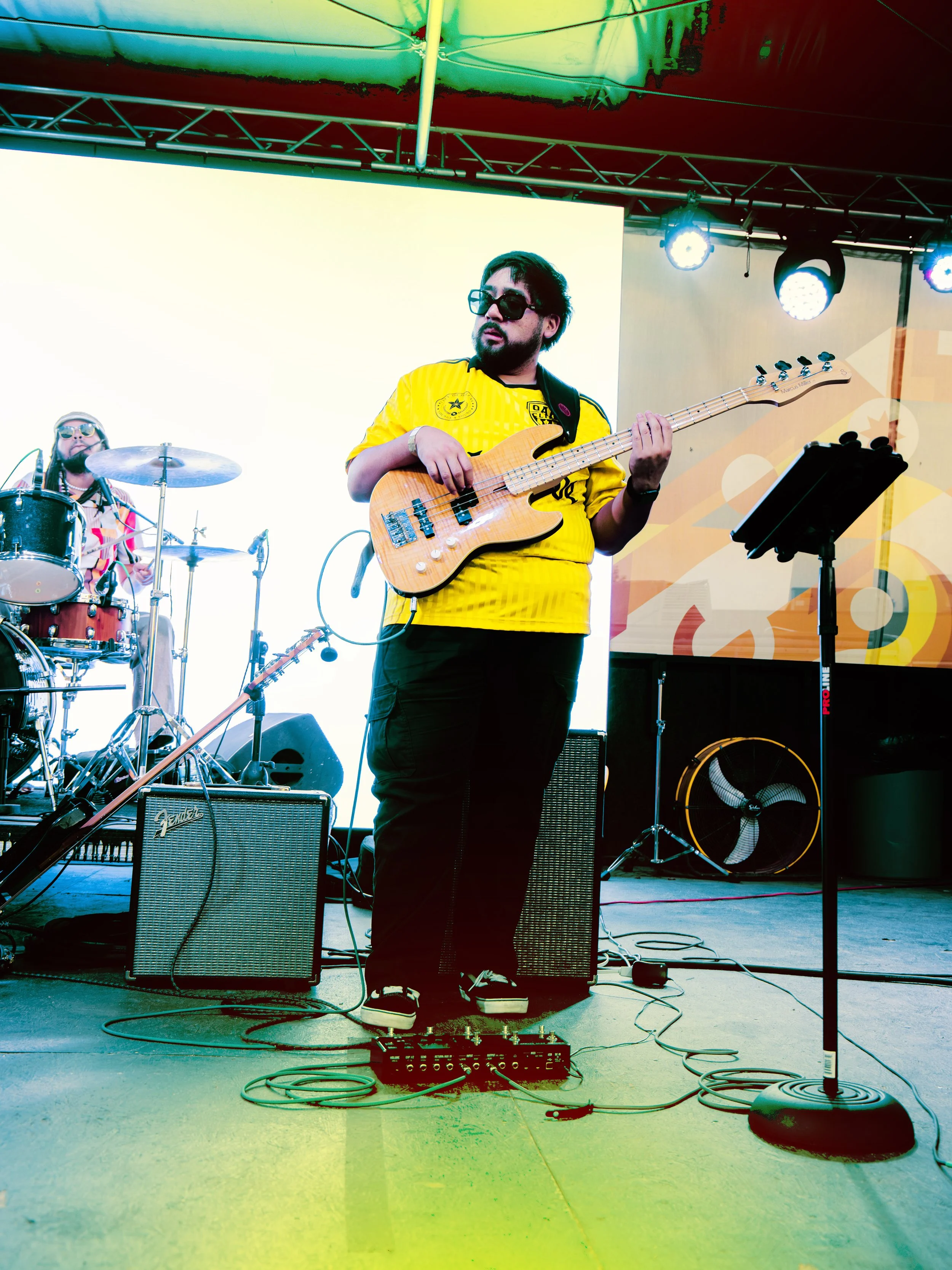 A musician playing a bass guitar on stage with a drummer in the background. The stage has colorful lighting and equipment including guitar amplifiers, microphone, music stand, and stage lights.