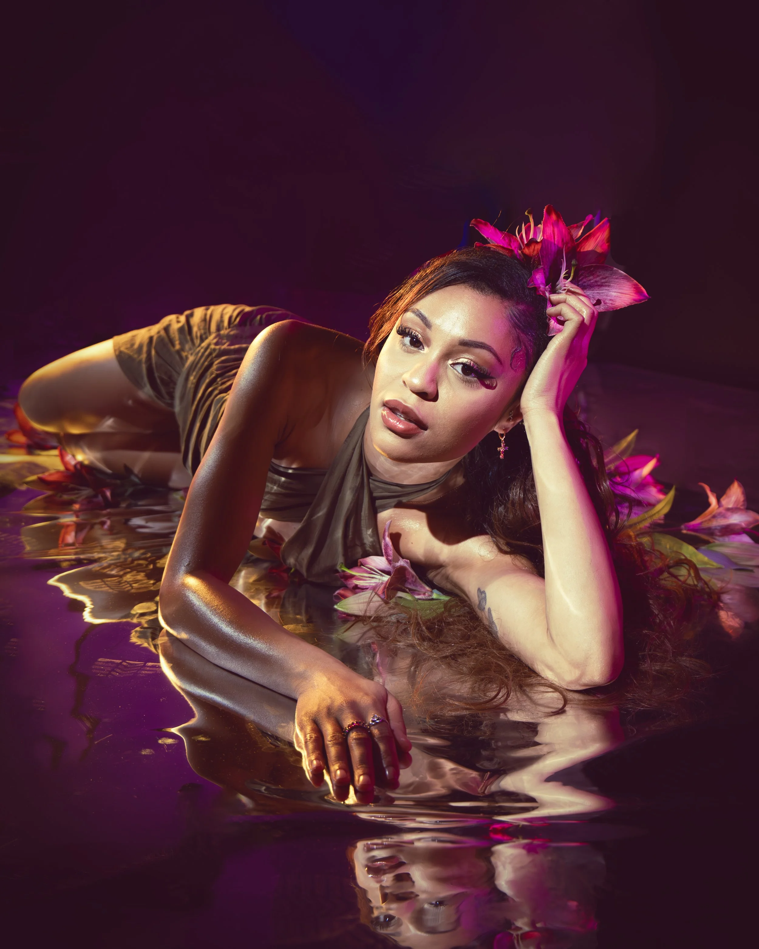 Local Dallas, Texas, DFW Non-traditional Fashion Editorial Creative Portraits Cover Art Photographer Photoshoot. Musician Rebelle Flor . A woman with long dark hair, wearing makeup and jewelry, lying on her side on water with pink flowers around her,