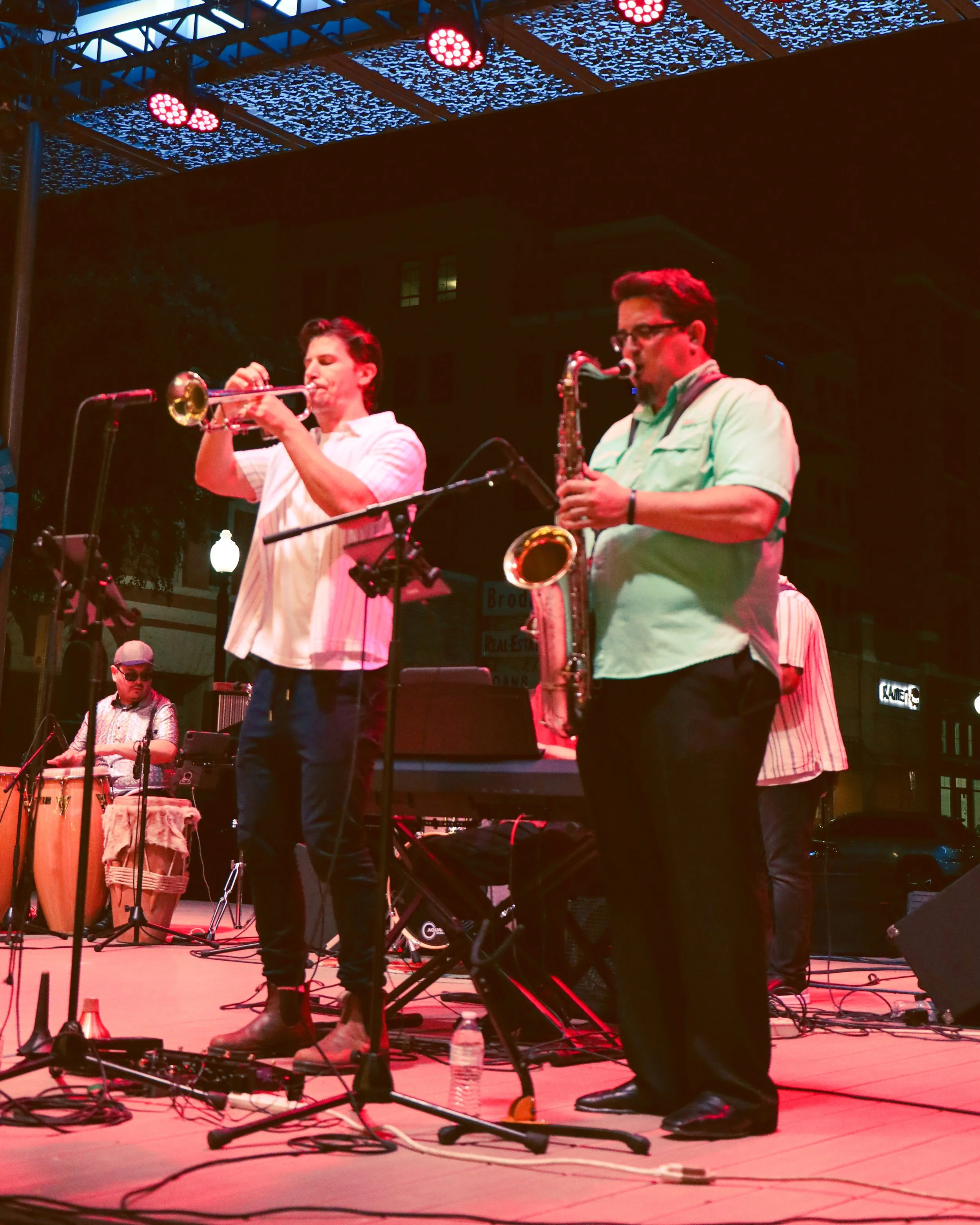 Musicians performing on a stage at night, with one playing a trumpet and another playing a saxophone, surrounded by various musical instruments and equipment.