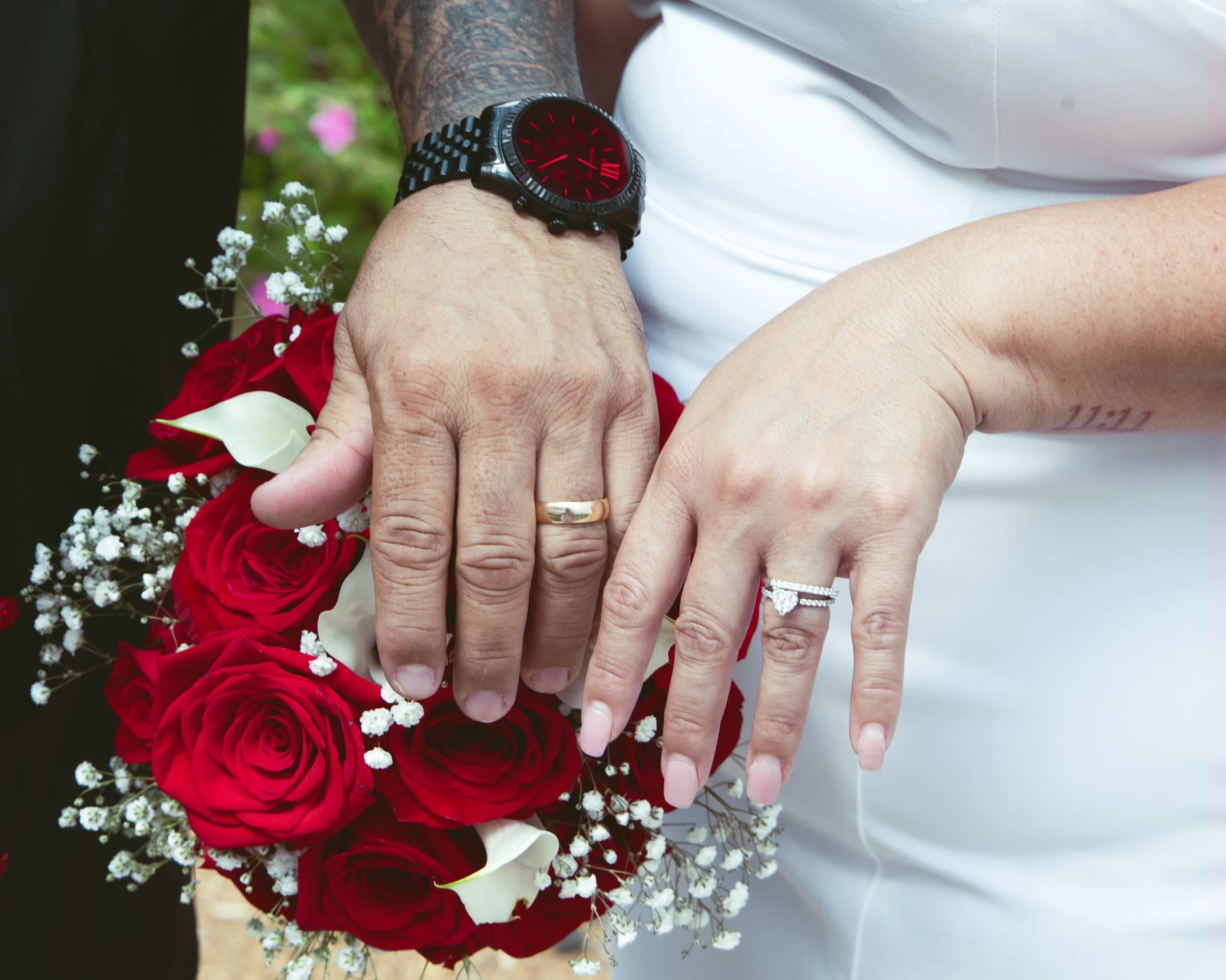 Close-up of a couple holding hands, with wedding rings visible on their fingers. The woman holds a bouquet of red roses and white flowers. The man wears a black wristwatch with a red face. The background is blurred.