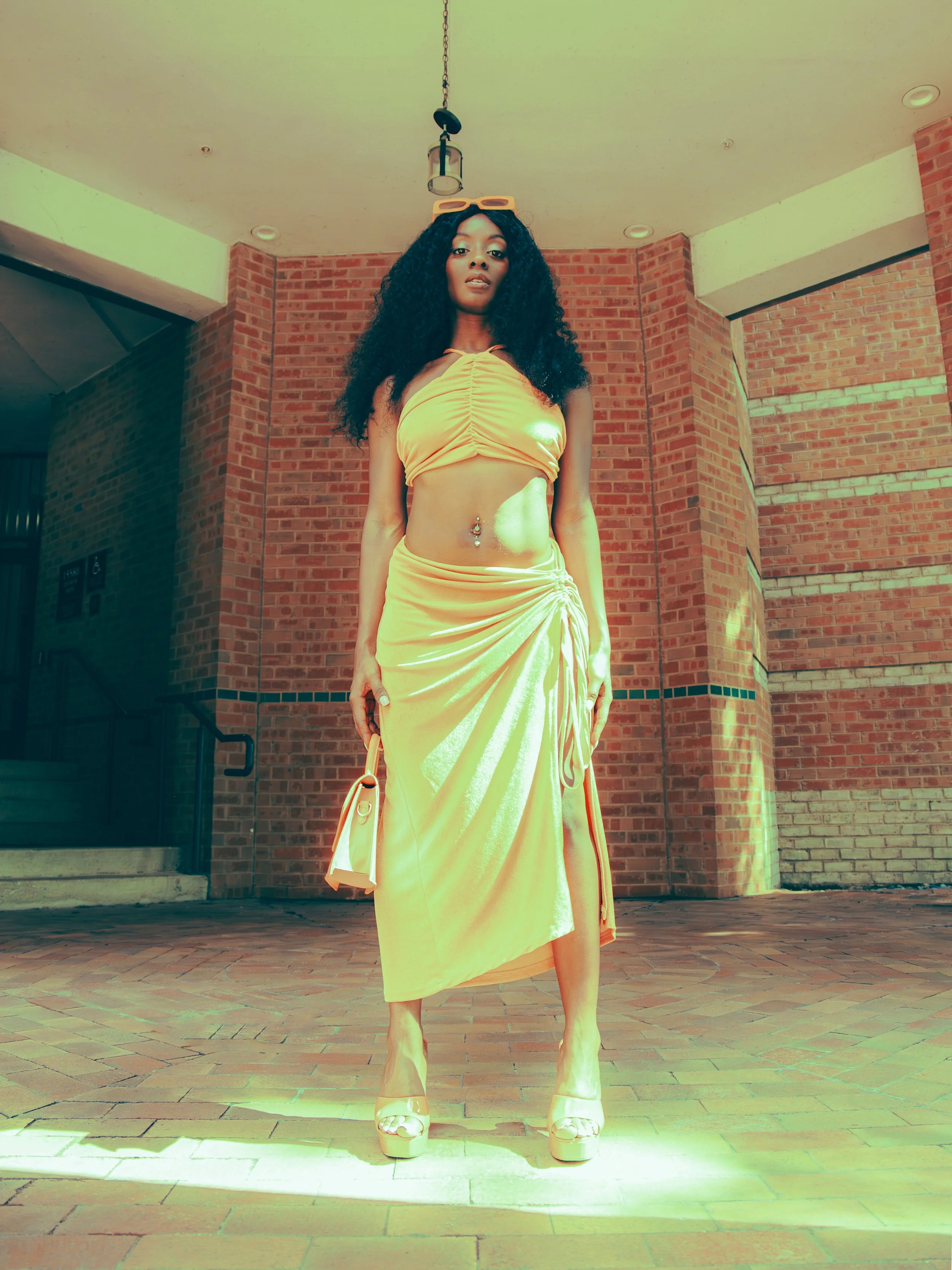 Local Dallas, Texas, DFW Non-traditional Fashion Editorial Creative Portraits Cover Art Photographer Photoshoot. A woman with long curly black hair wearing a yellow crop top, a yellow skirt, yellow sunglasses, and high heels standing in front of a br