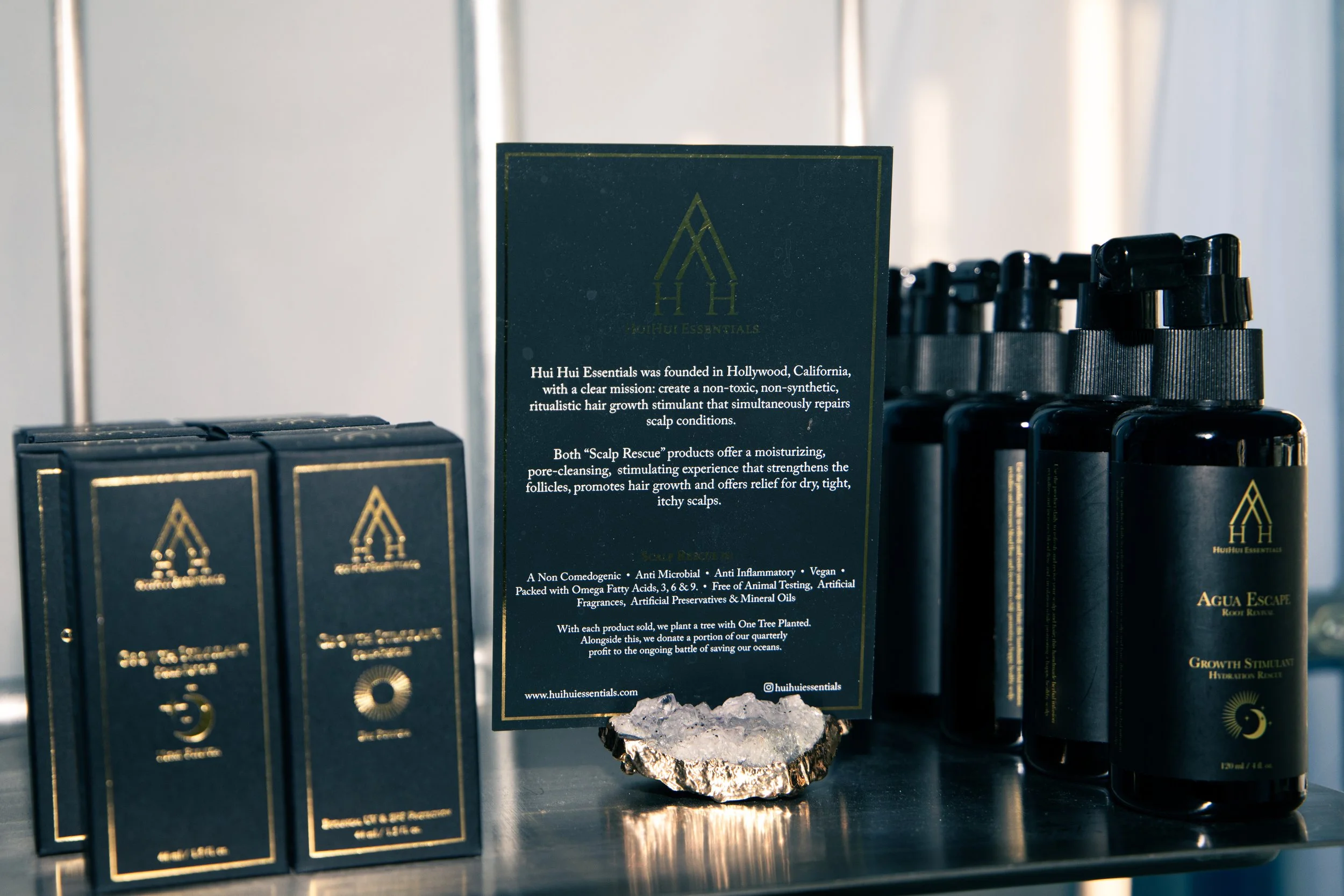 Display of Hui Hui Essentials hair care products on a shelf, with a sign explaining the company's mission and benefits, set against a minimalist backdrop.