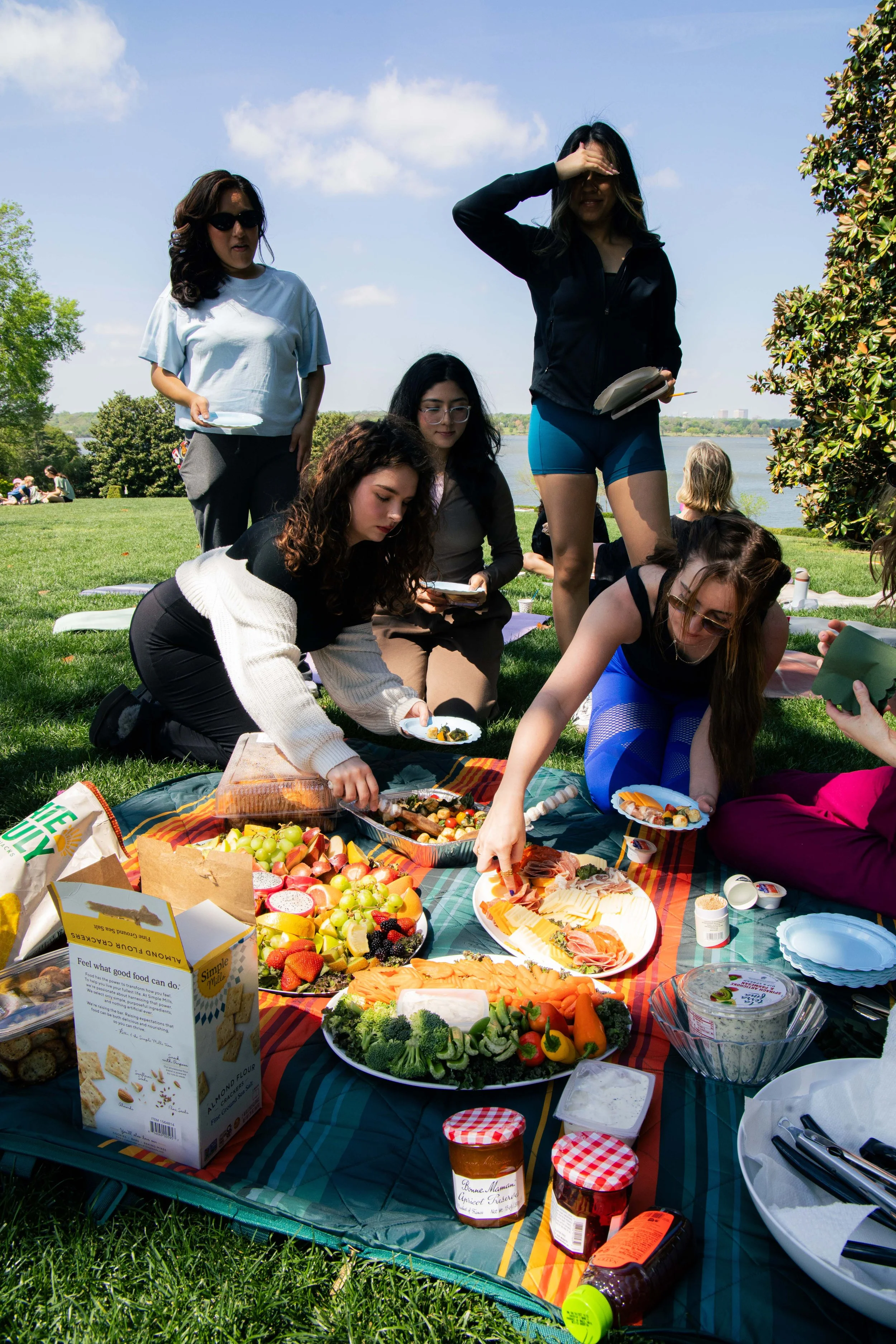 A group of women having a picnic outdoors by a lake with food spread on a blanket, including fruits, vegetables, cheese, and snacks.
