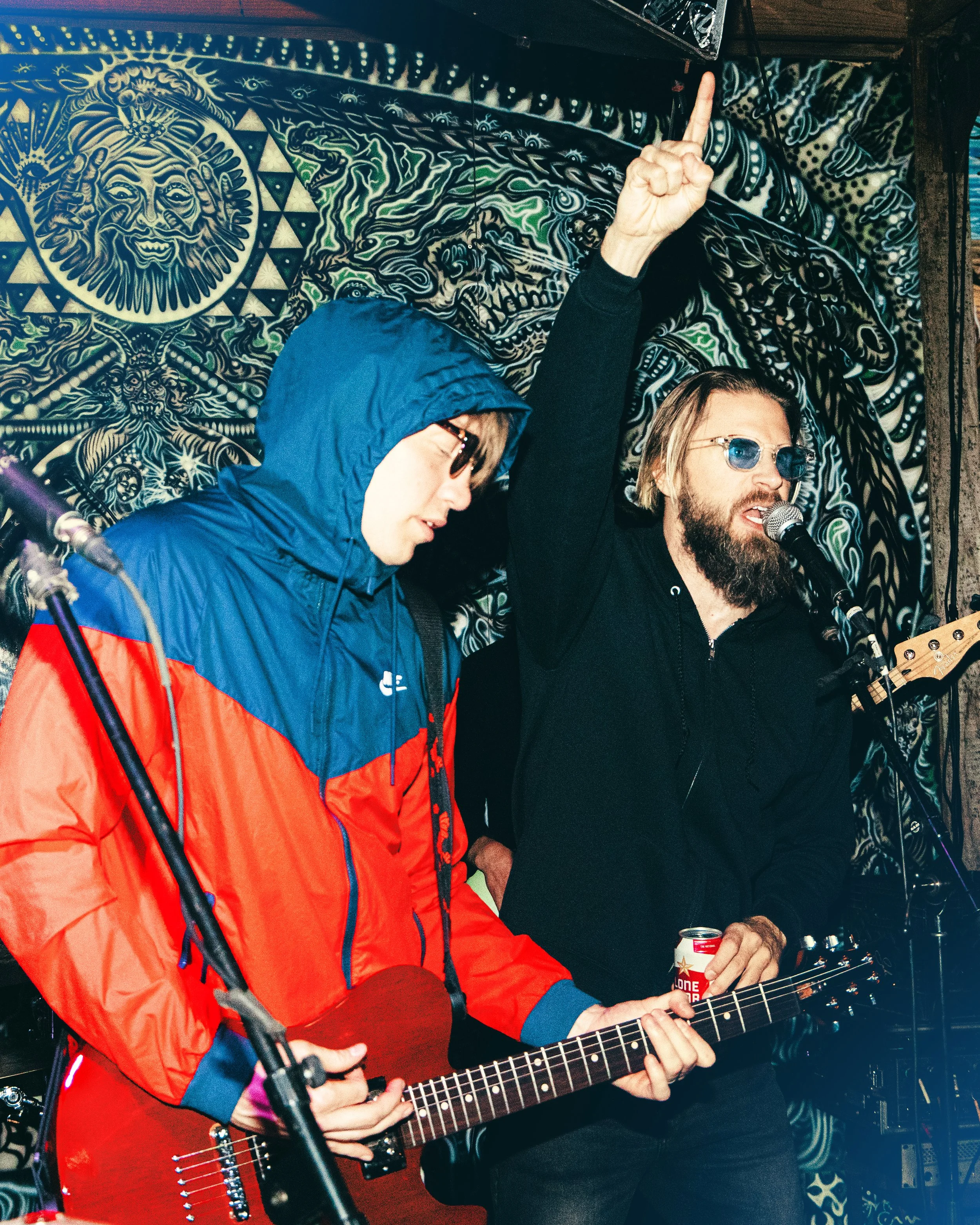 Two musicians performing at a venue with a colorful, psychedelic tapestry background. One person wearing sunglasses and a black hoodie is playing a guitar and singing into a microphone, while the other person in a blue and red jacket, also wearing su
