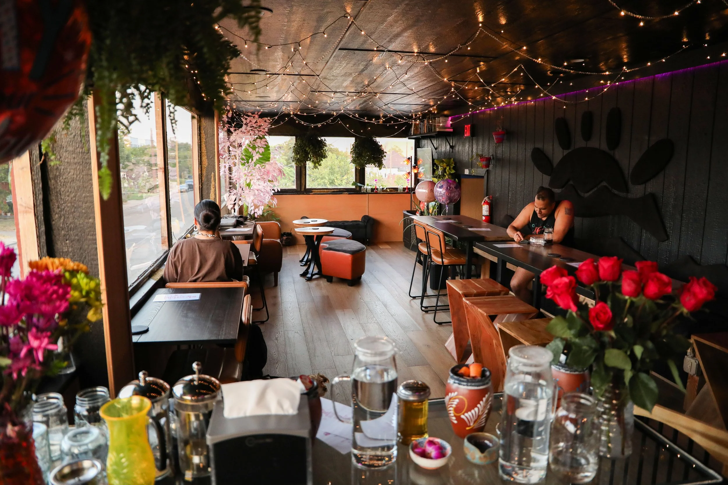 Inside a cozy, decorated cafe with string lights on the ceiling, large windows letting in natural light, and potted plants. Two people are seated, one facing away and the other looking down, surrounded by tables with drinks, flowers, and decorative i