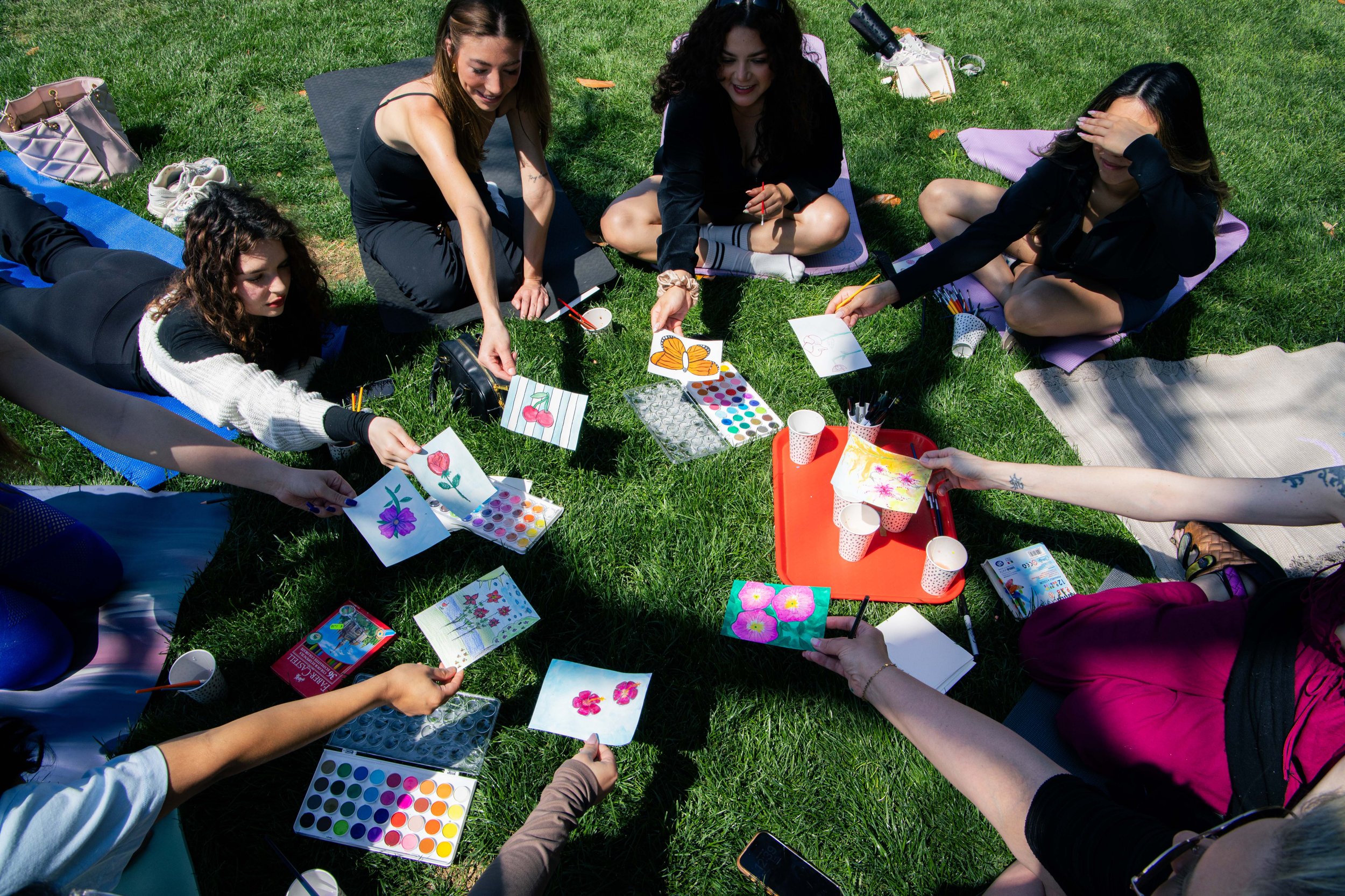 A group of women sitting on the grass during a sunny day, engaging in watercolor painting and art activities, with art supplies and bags around them.