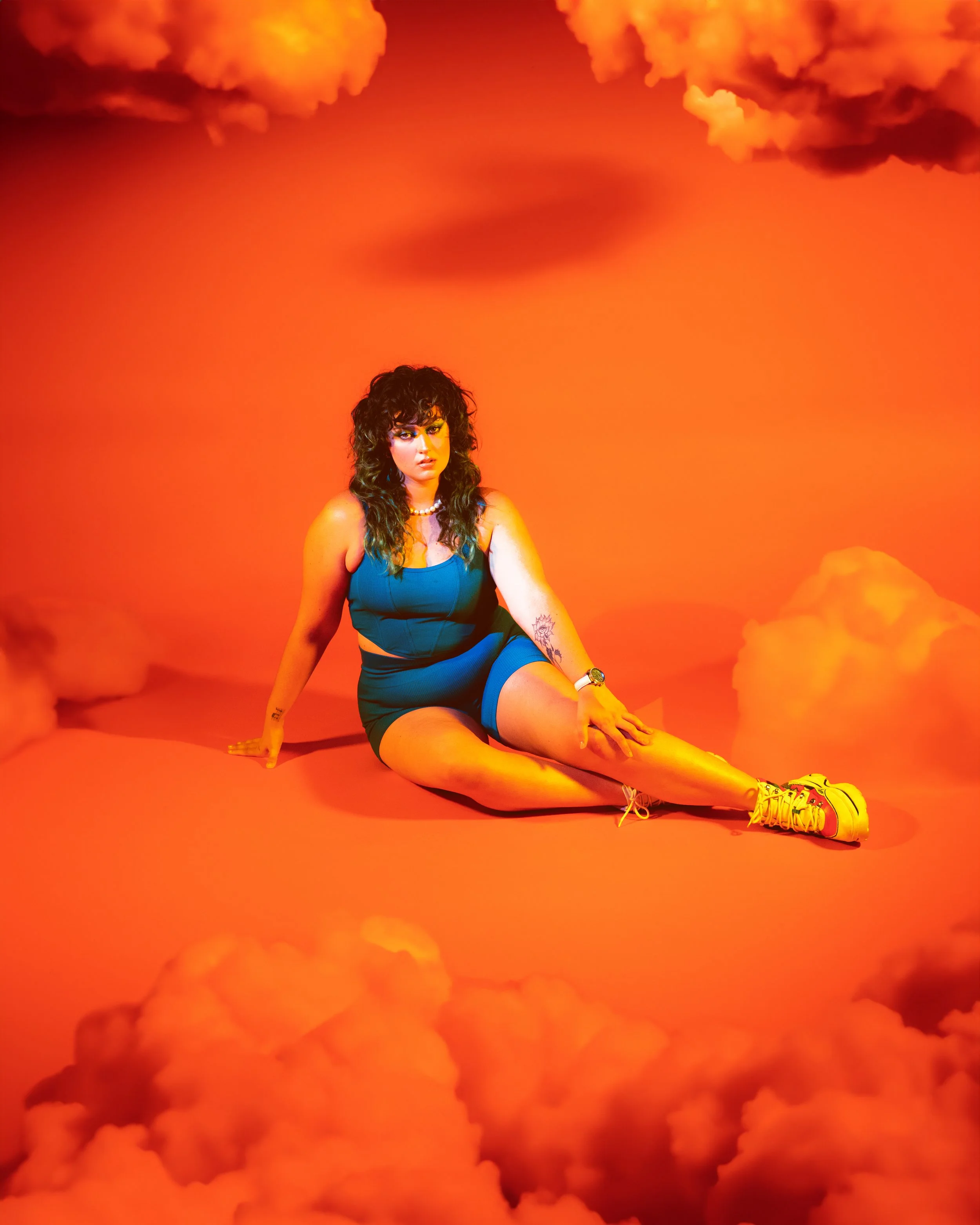 Local Dallas, Texas, DFW Non-traditional Fashion Editorial Creative Portraits Cover Art Photographer Photoshoot. @roseypears . A woman with dark curly hair wearing a blue tank top and shorts, sitting on an orange floor with a bright orange background