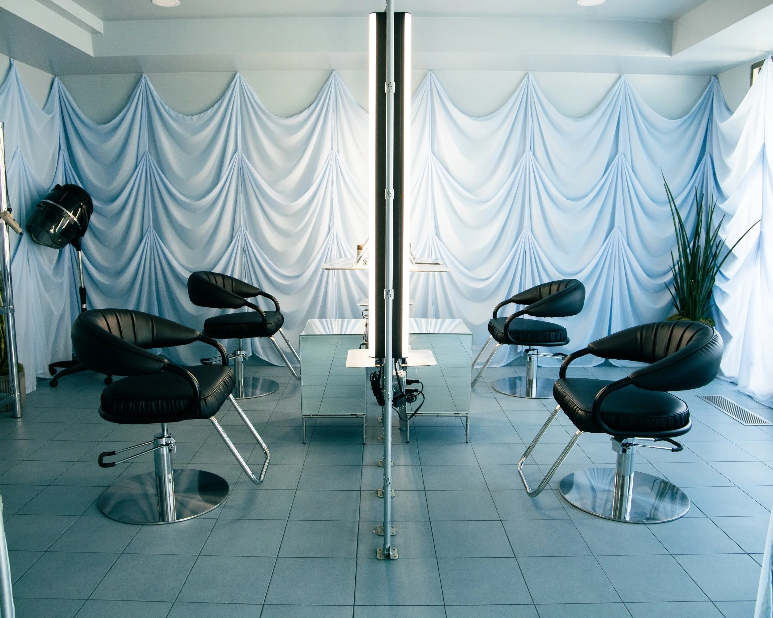 Modern indoor room with a white fabric backdrop, three black chairs with metal bases, a standing studio light, and a mirror in the center reflecting the scene. There are also some green plants visible on the right side.