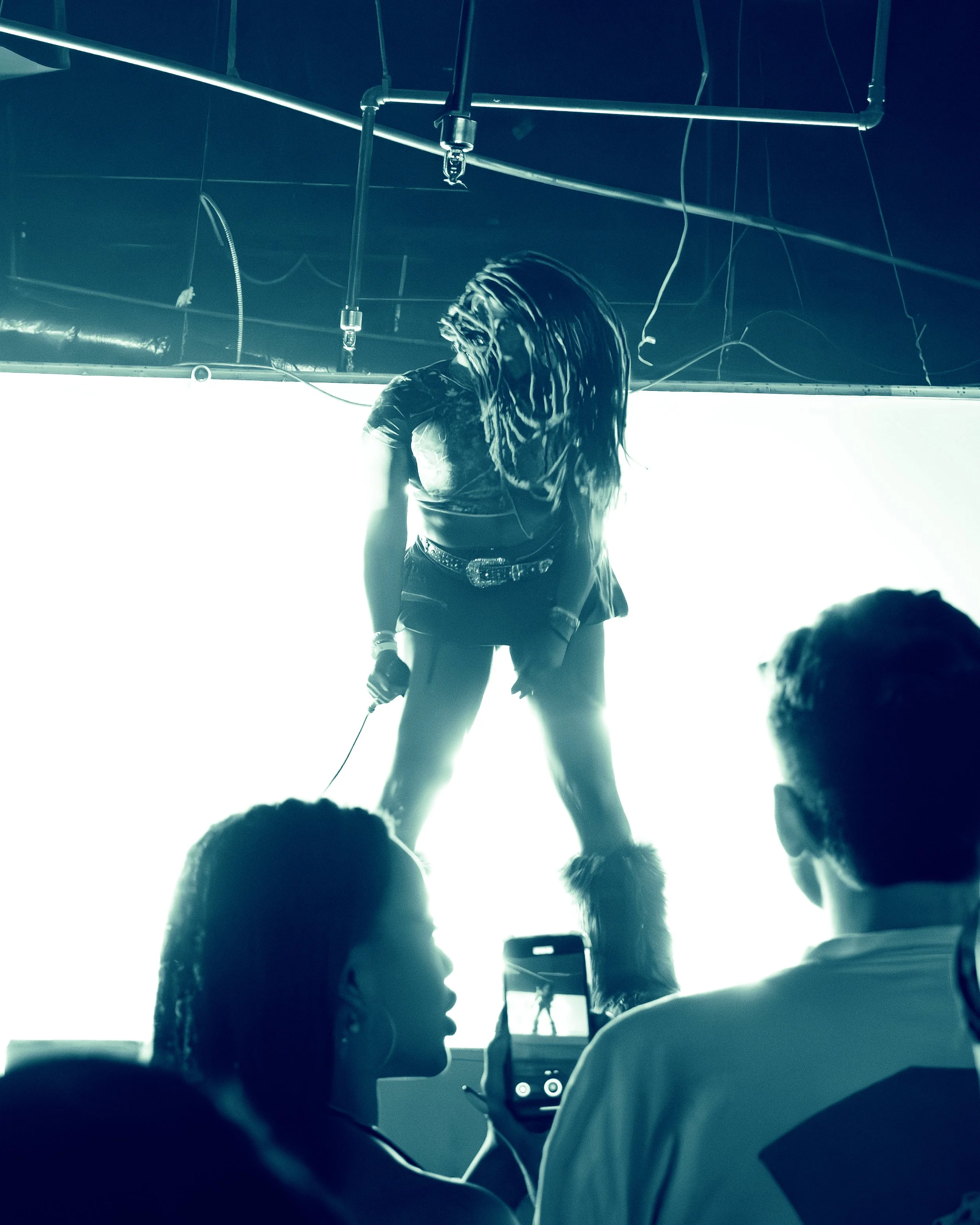 People seated in a dark theater or auditorium, watching a person dancing or performing on stage under bright lighting. The performer has long, braided hair and is wearing casual clothing.