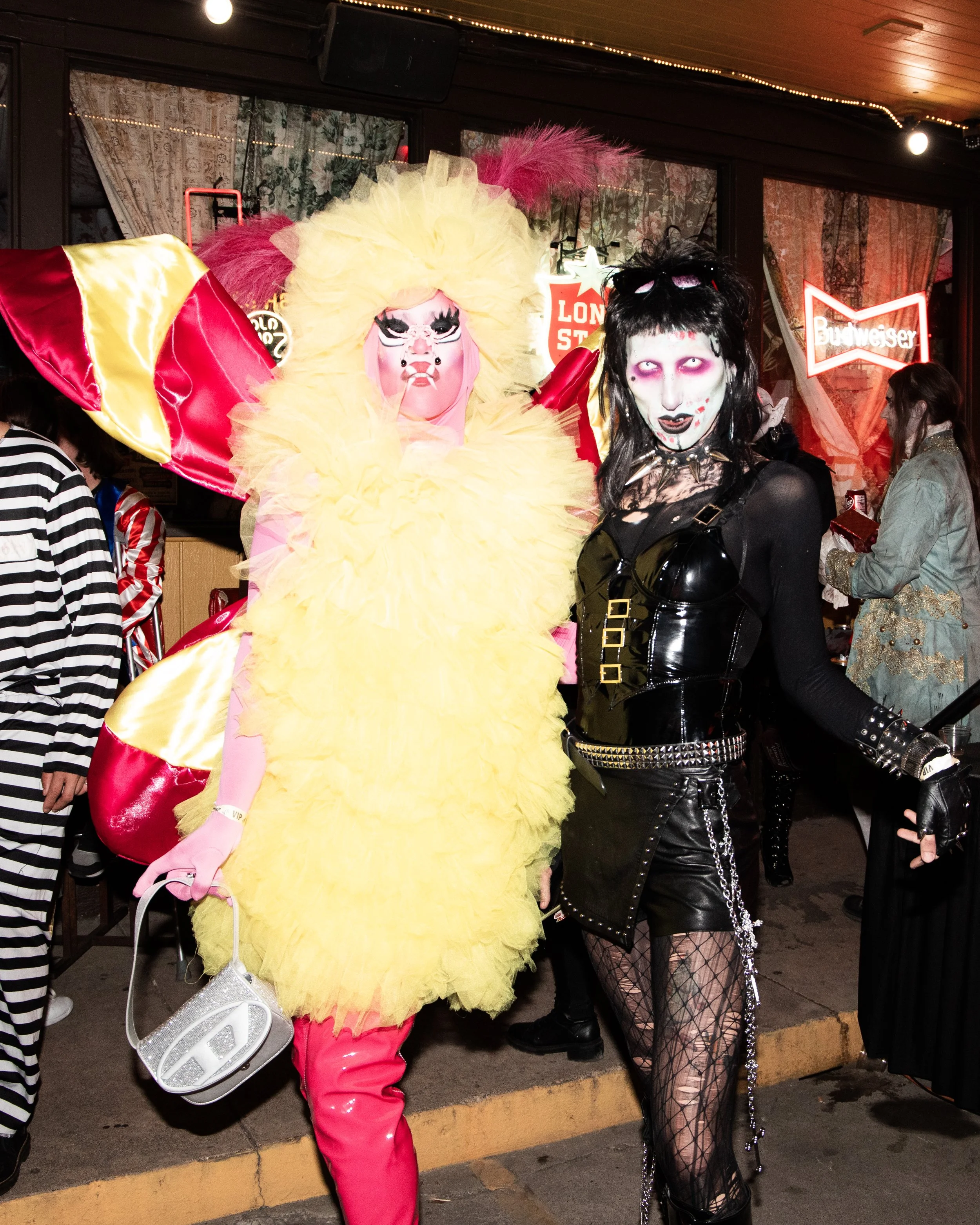 People in costumes at a nighttime event, including a person in a pink and yellow feathered costume with a mask and another person in gothic makeup with fishnet stockings and black clothing. Neon signs and decorated curtains are in the background.