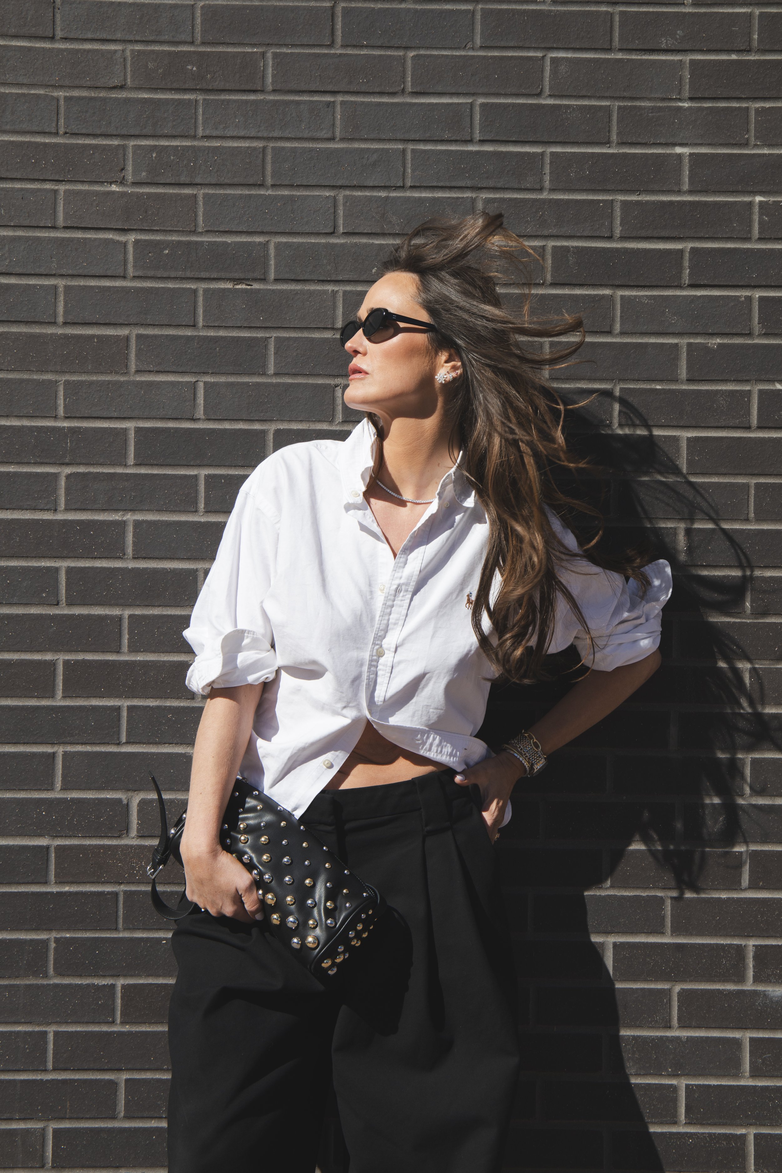 Local Dallas, Texas, DFW Non-traditional Fashion Editorial Creative Portraits Cover Art Photographer Photoshoot. Revel Workshop . A woman with long, wavy hair wearing dark sunglasses, a white button-up shirt tied at the waist, and black pants, standi