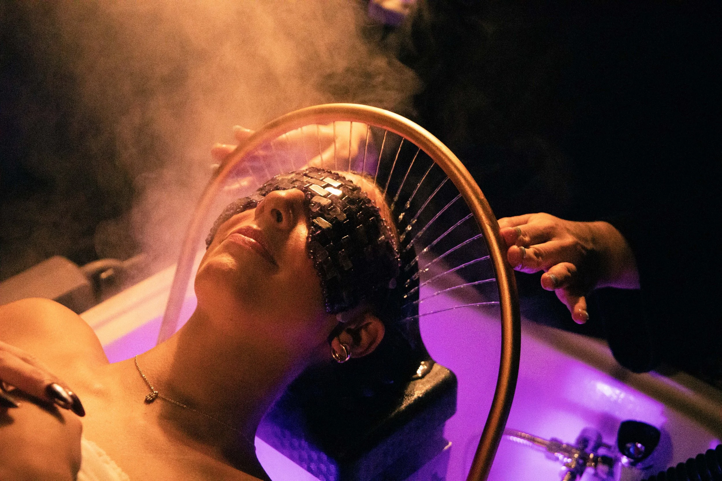A person with short dark hair and earrings is reclining with a bicycle wheel on their face, surrounded by colorful purple and pink lighting, and smoke or vapor in the background.