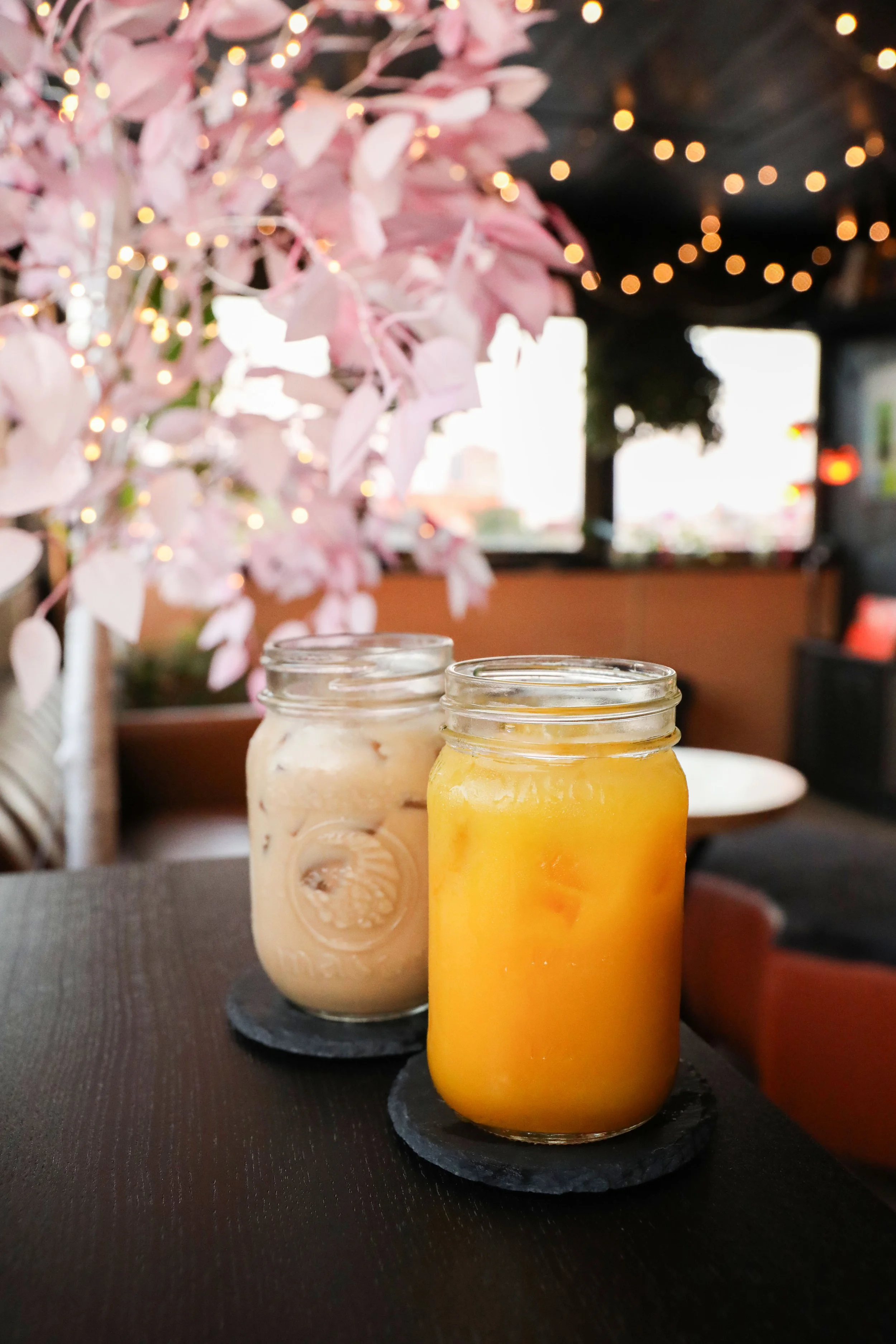 Two mason jars filled with creamy yellow and orange smoothies on a black slate coaster, on a black placemat, in front of pink flowering branches with fairy lights and blurred background.