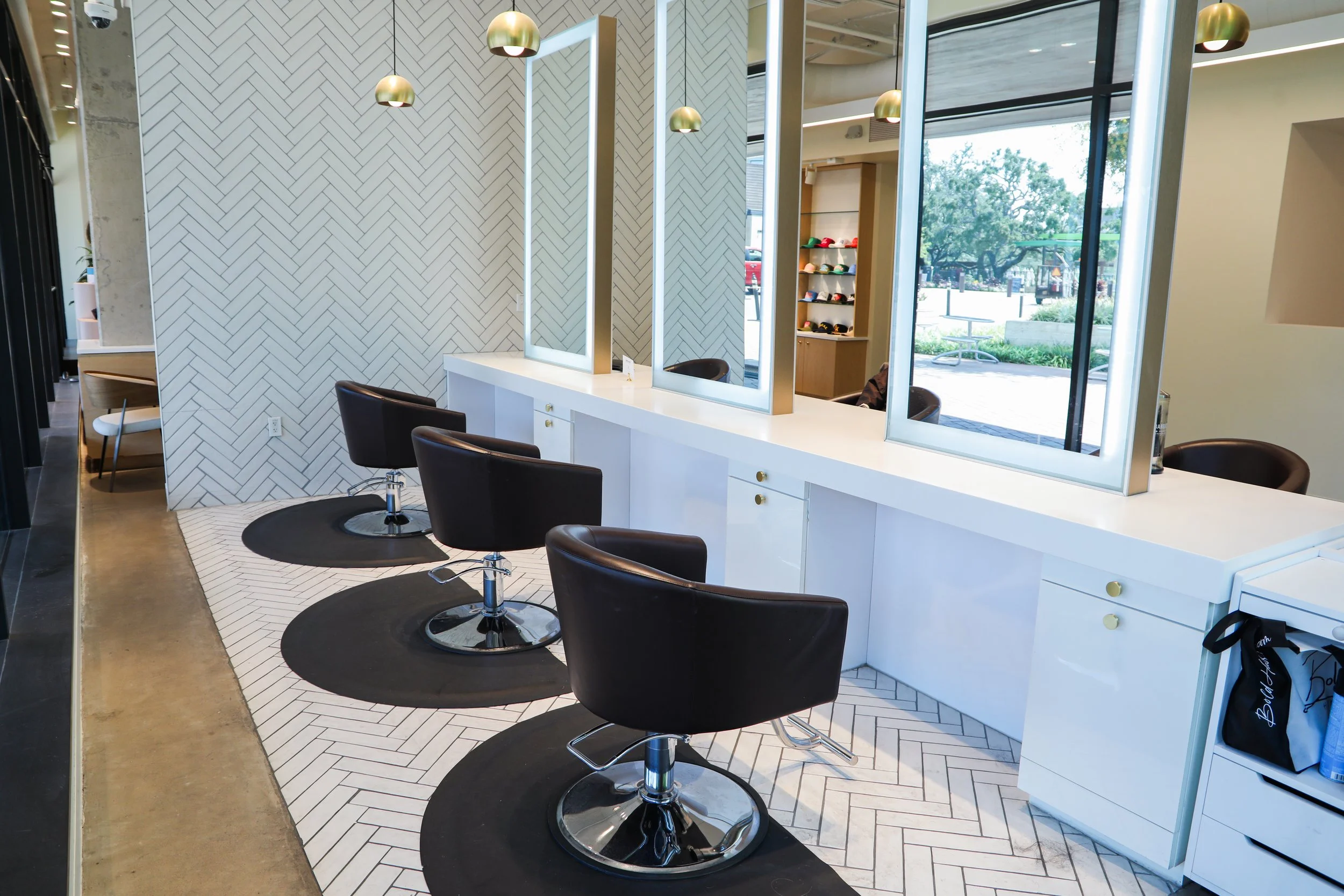 Inside a modern hair salon with a white counter, black salon chairs, large mirrors, and a window showing greenery outside.