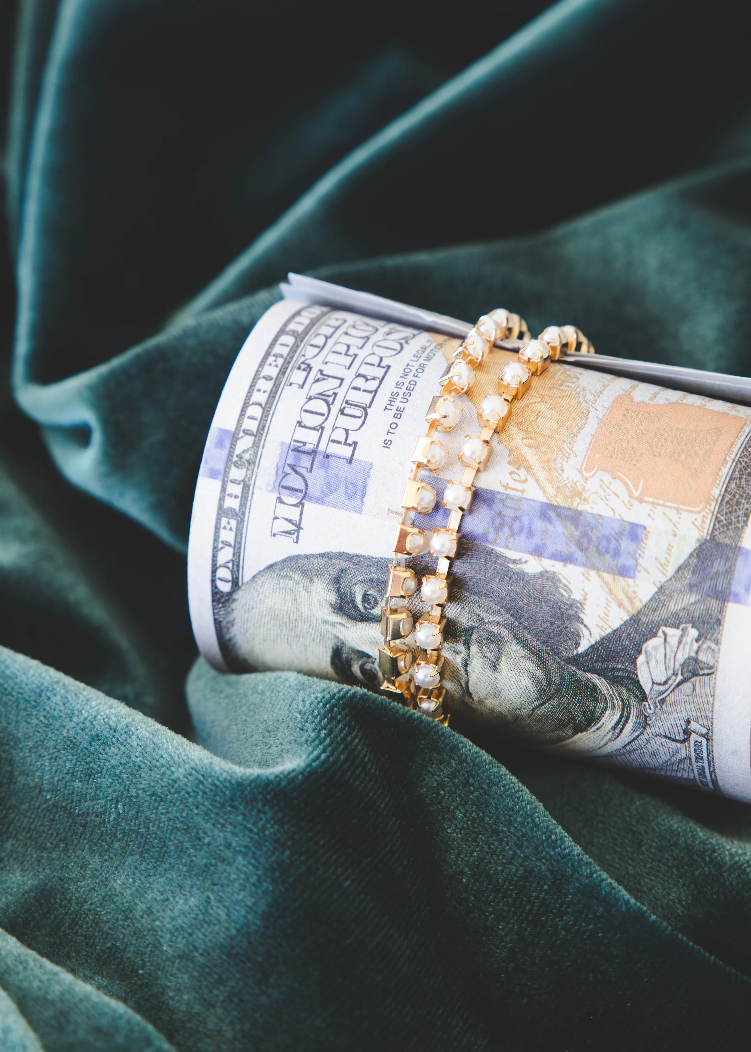 A twisted U-shaped gold bracelet with white pearls on top of a rolled American hundred-dollar bill, which is resting on dark green velvet fabric.