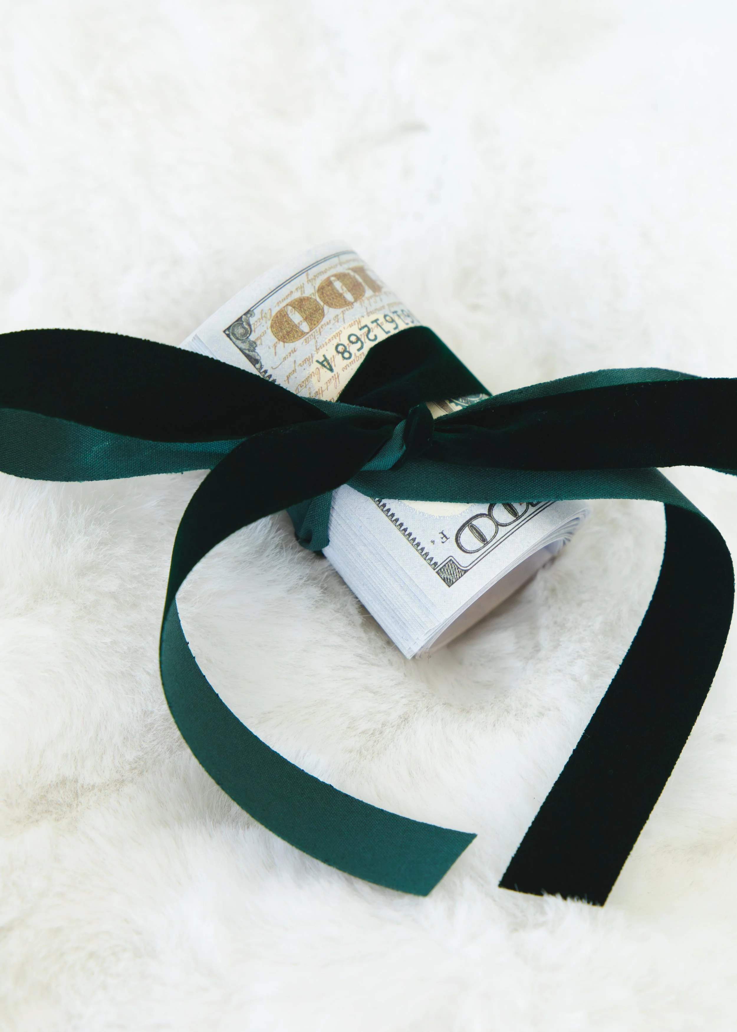 Rolled bundle of a 100-dollar bill tied with a dark green ribbon, resting on a white furry surface.