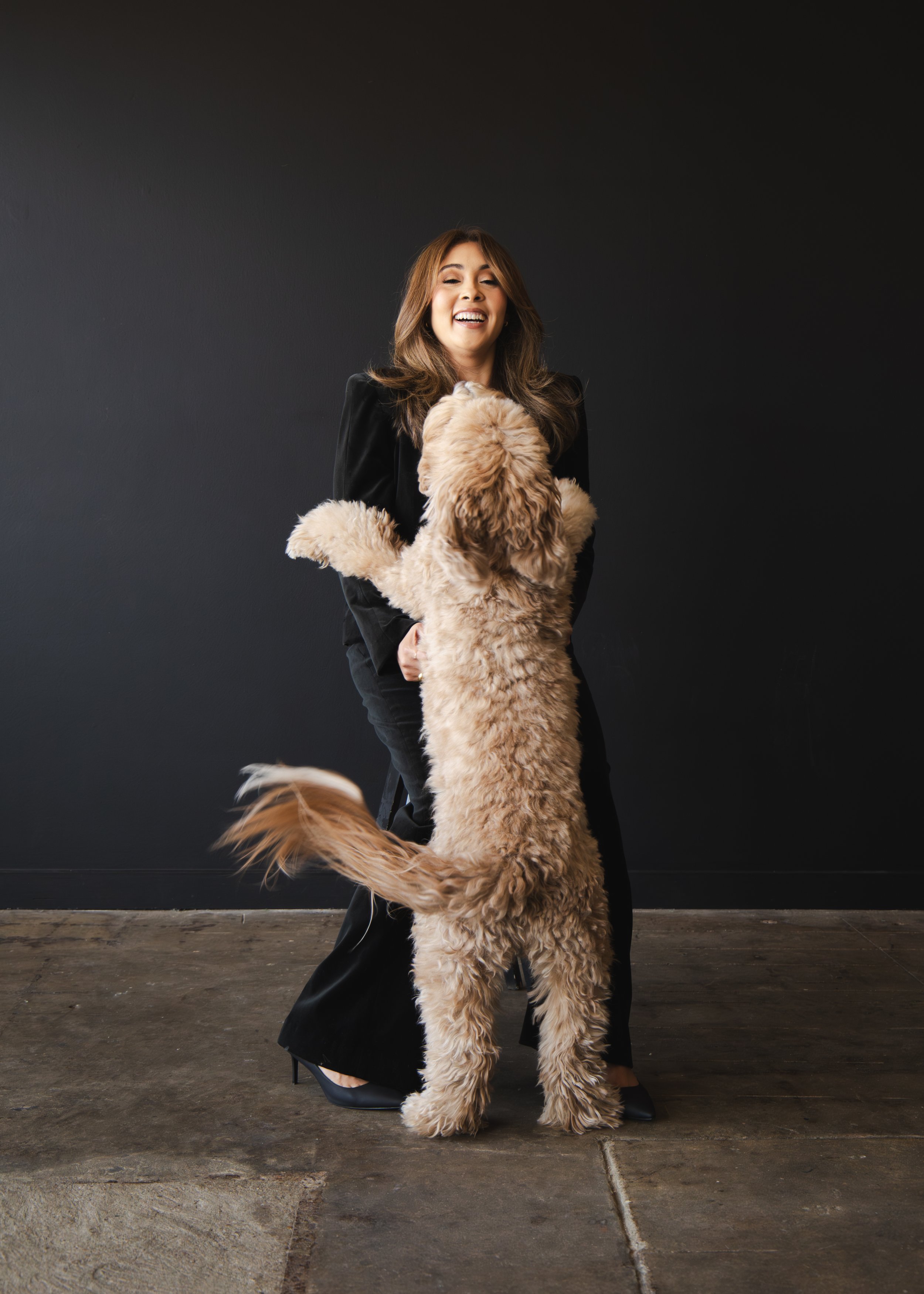 Local Dallas, Texas, DFW Non-traditional Fashion Editorial Creative Portraits Cover Art Photographer Photoshoot. Adriana Martinez , Revel Workshop , Vida Wealth Club . A woman with long brown hair, smiling, wearing a black outfit, standing on a woode