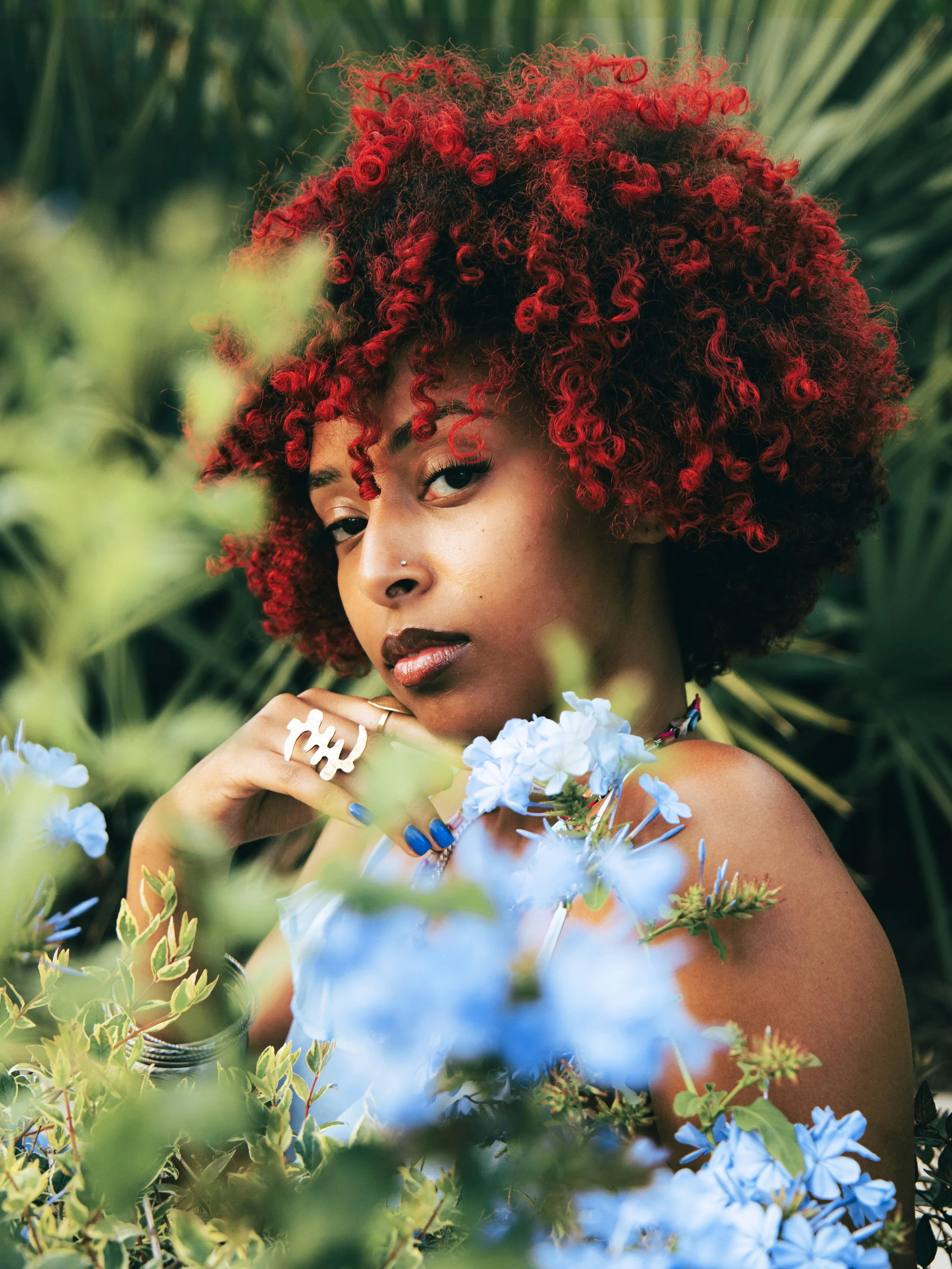 Local Dallas, Texas, DFW Non-traditional Fashion Editorial Creative Portraits Cover Art Photographer Photoshoot. Musician Aden , A woman with vibrant, curly red hair and a nose piercing posing among green plants and blue flowers.