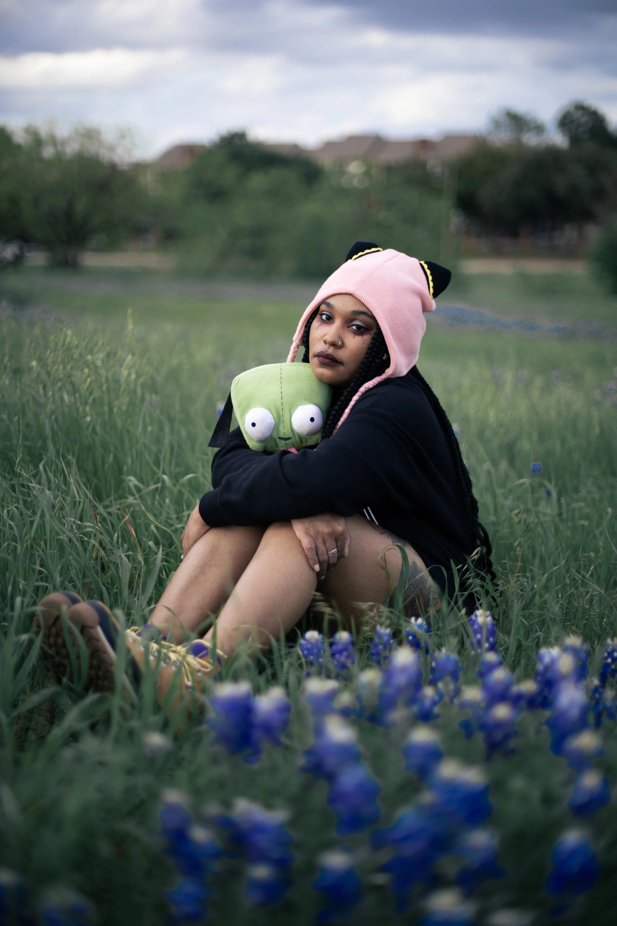 Local Dallas, Texas, DFW Non-traditional Editorial Creative Portraits Cover Art Photographer. Bluebonnets. Bluebonnet photoshoot. Texas Bluebonnets. Musician Igimeji . A woman with braided hair, wearing a pink cat-eared hat, dark lipstick, and a blac