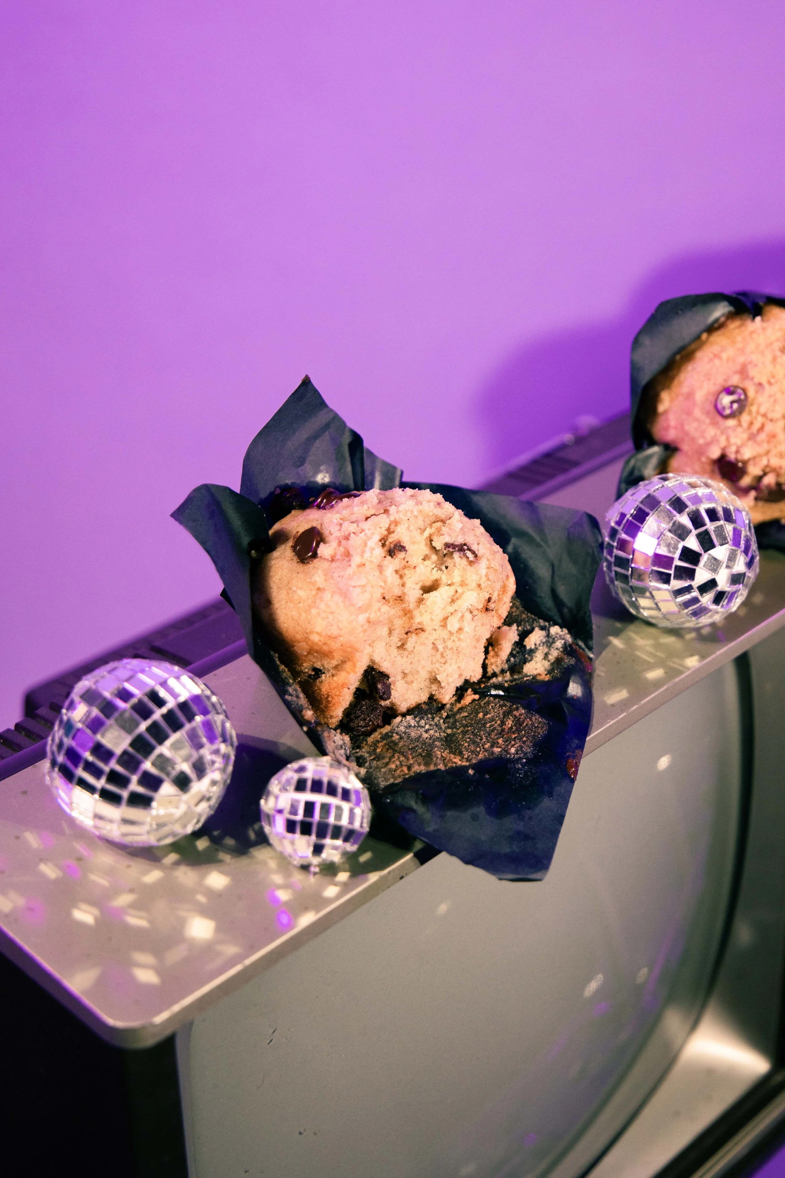Chocolate chip cookies wrapped in black paper with mini disco balls around them, placed on a silver reflective surface with purple lighting.