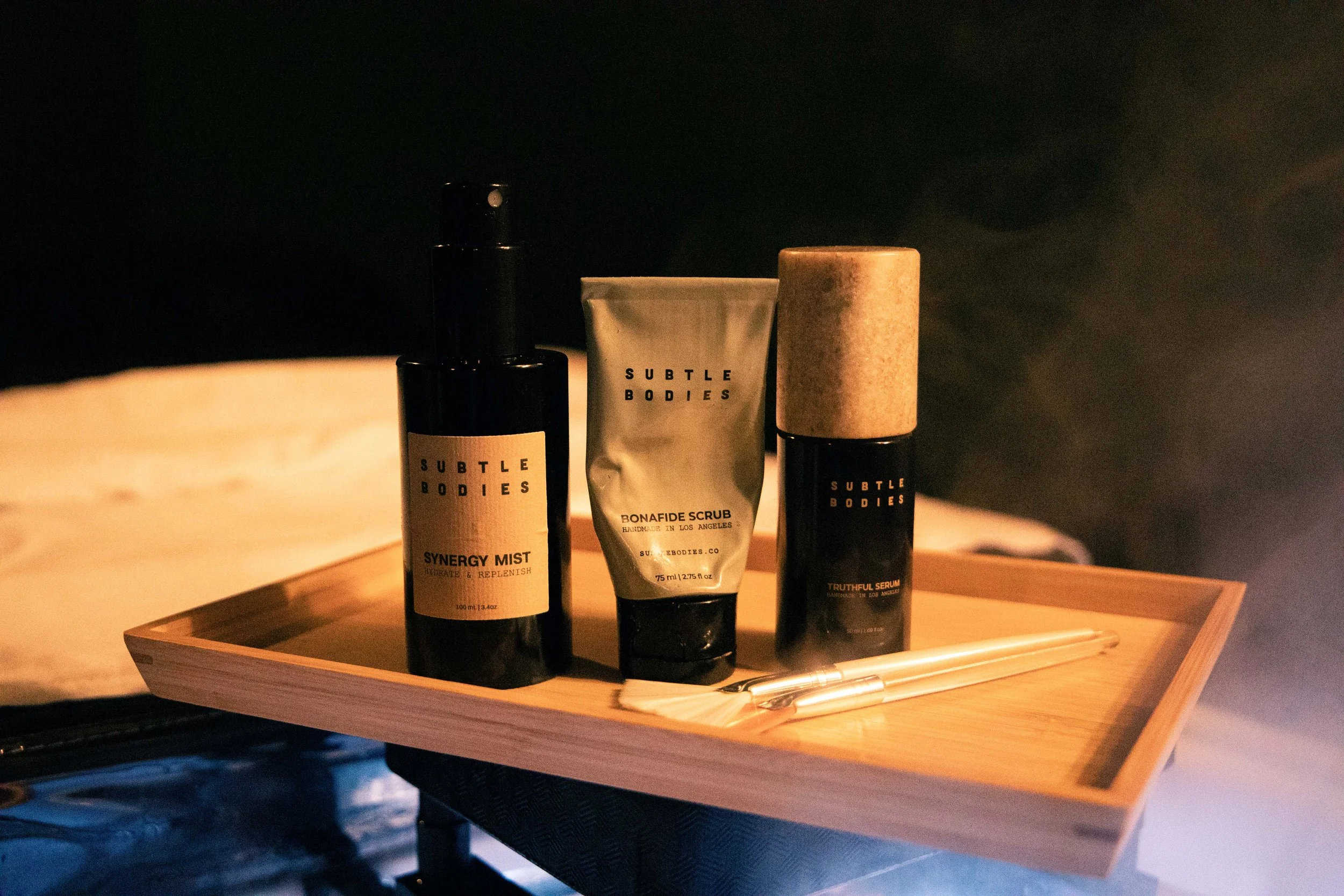 A wooden tray holding three skincare products and a pen on a wooden surface, with a dark background.