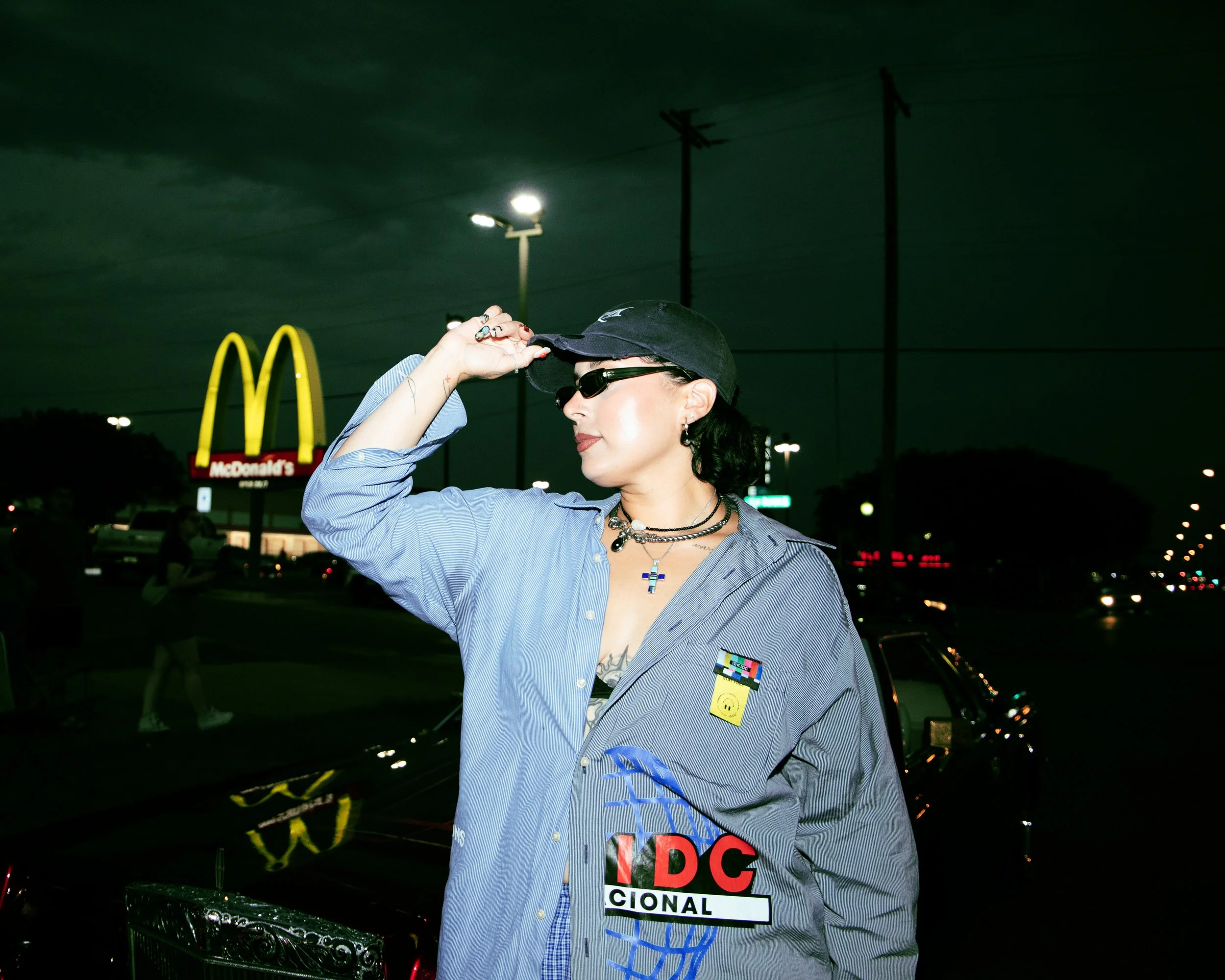 Local Dallas, Texas, DFW Non-traditional Fashion Editorial Creative Portraits Cover Art Photographer Photoshoot. Nataly Medina , Crecer Dallas . A woman adjusts her black baseball cap and sunglasses in a parking lot at night. She wears layered neckla
