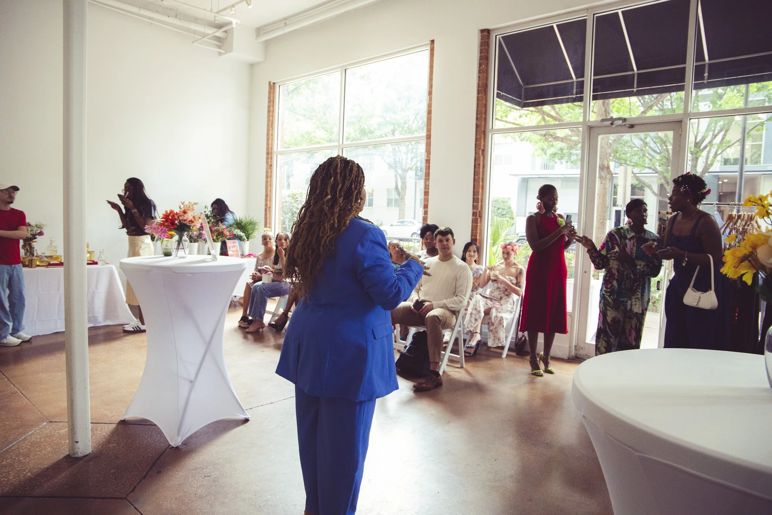 People gathered inside a well-lit room with large windows, some seated and some standing, engaged in conversations. There are flowers and decorative tables around, suggesting a social event or gathering.