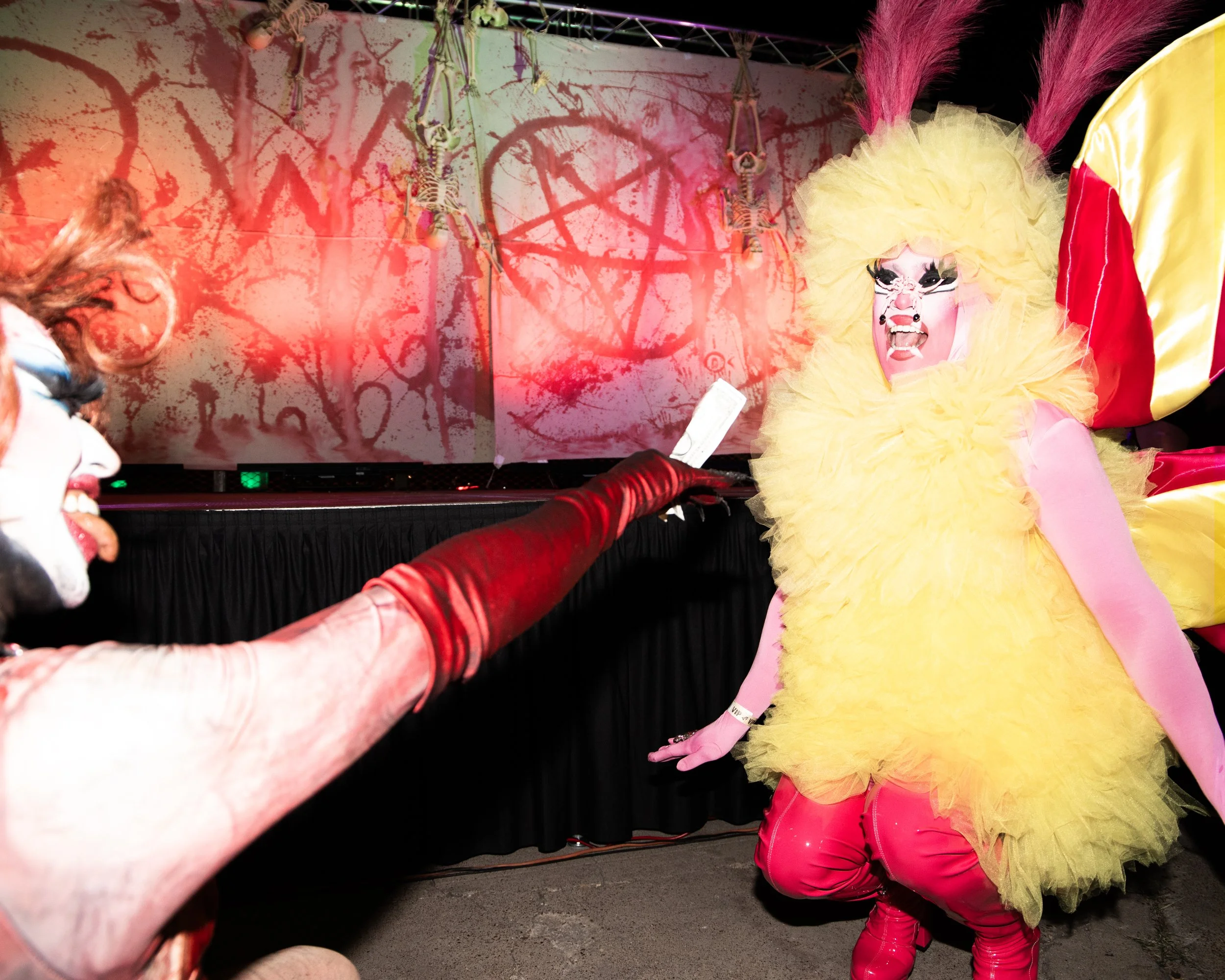 A performer dressed as a colorful clown or mime with a yellow fluffy costume, pink hair, and painted face, on stage with skeleton decorations and red paint splatters in the background.