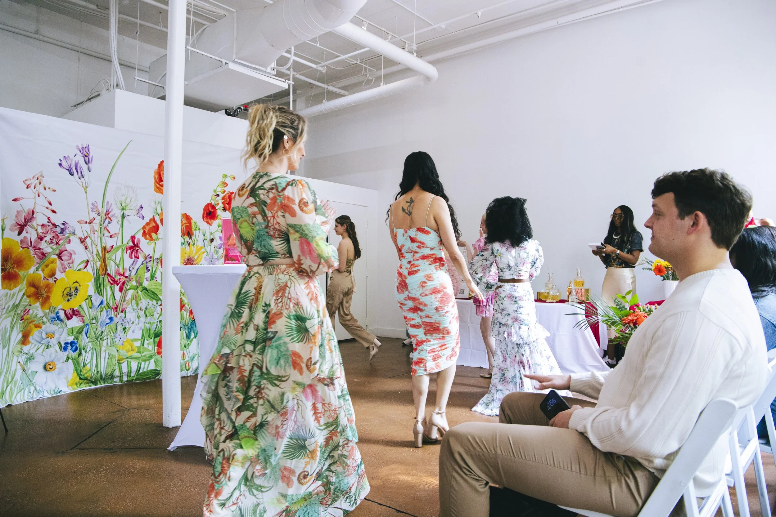 People gather in a bright indoor space with colorful floral decor, some standing and others sitting, enjoying a social event or gathering.