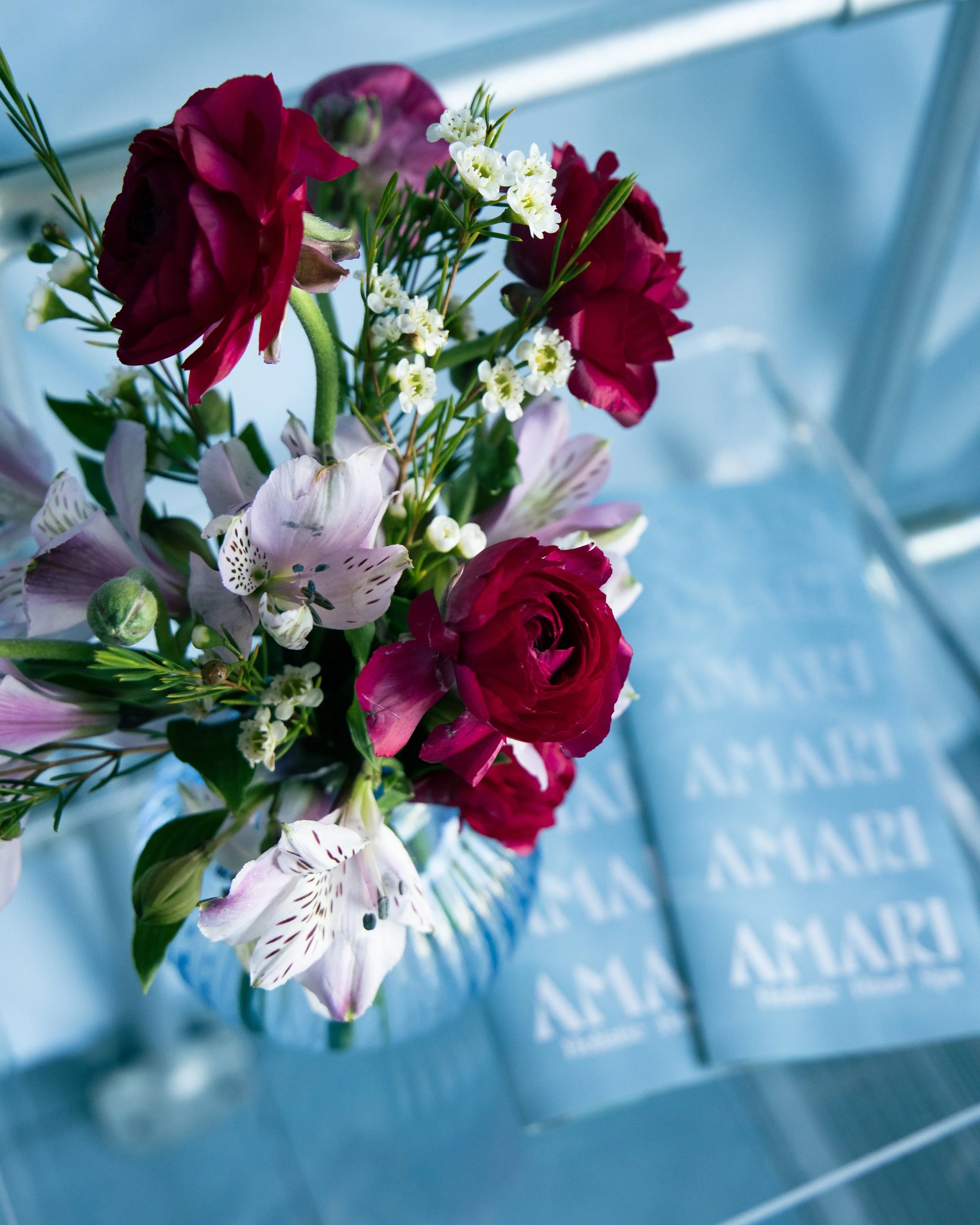 Close-up of a bouquet of mixed flowers including pink lilies, deep red roses, and small white flowers, set against a blue background with some text partially visible.