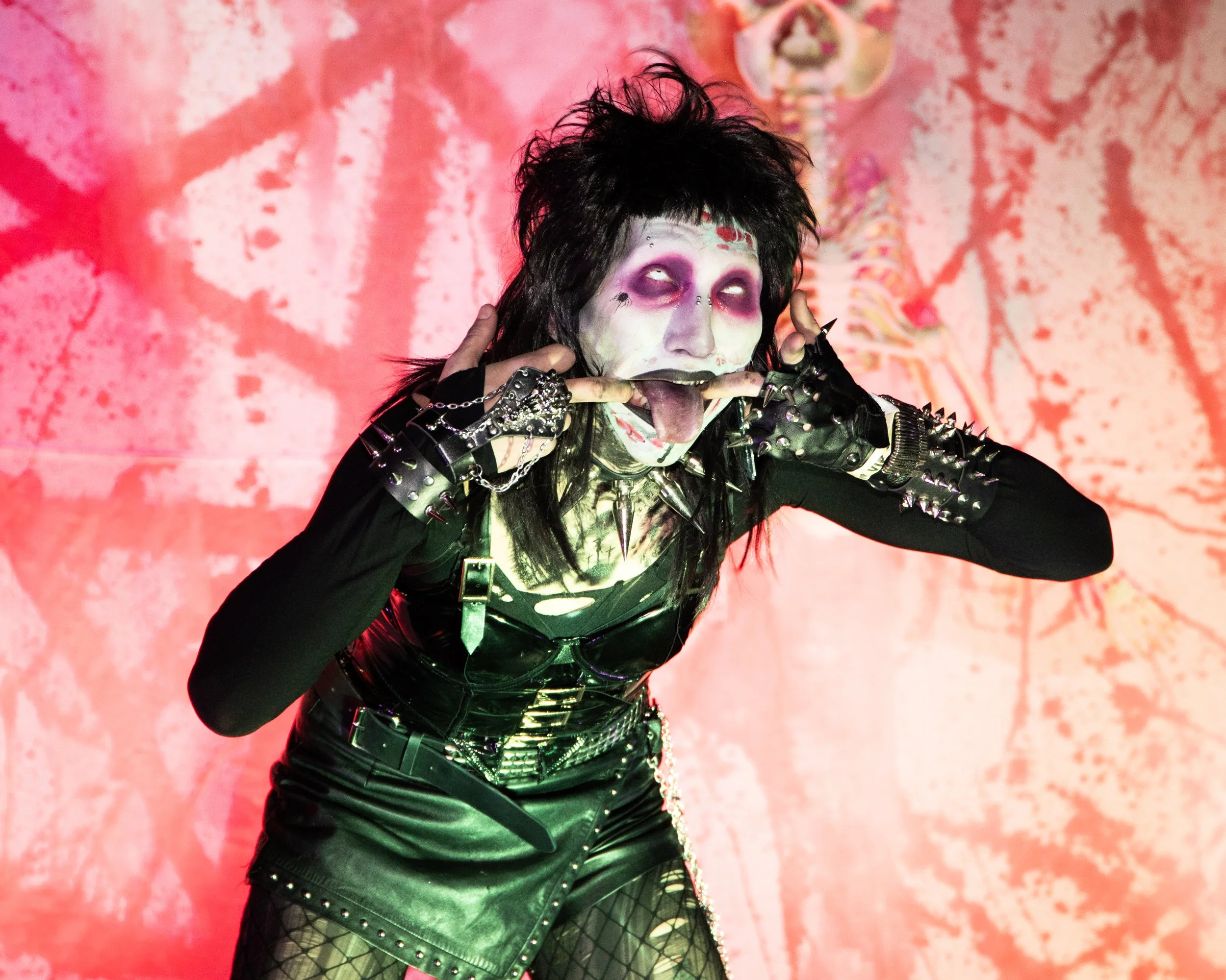 A person with dark, spiky hair and pale makeup, wearing a black leather outfit with spikes and chains, posing with fingers on their temples, sticking out their tongue, in front of a pink and red textured background.