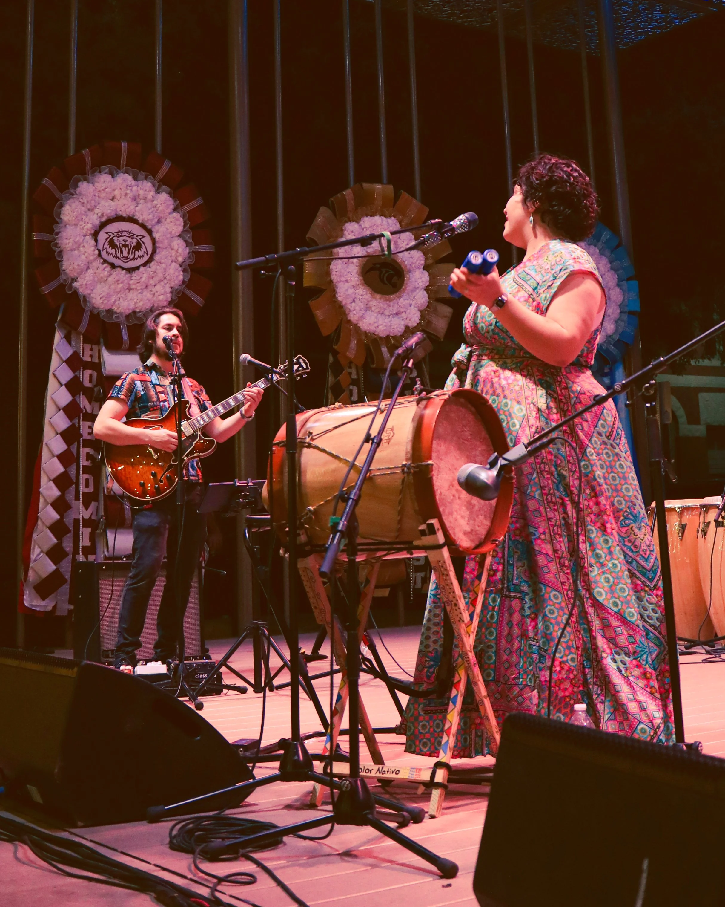Musicians performing on stage with floral wreaths and traditional instruments, including drums and guitars, under stage lighting.