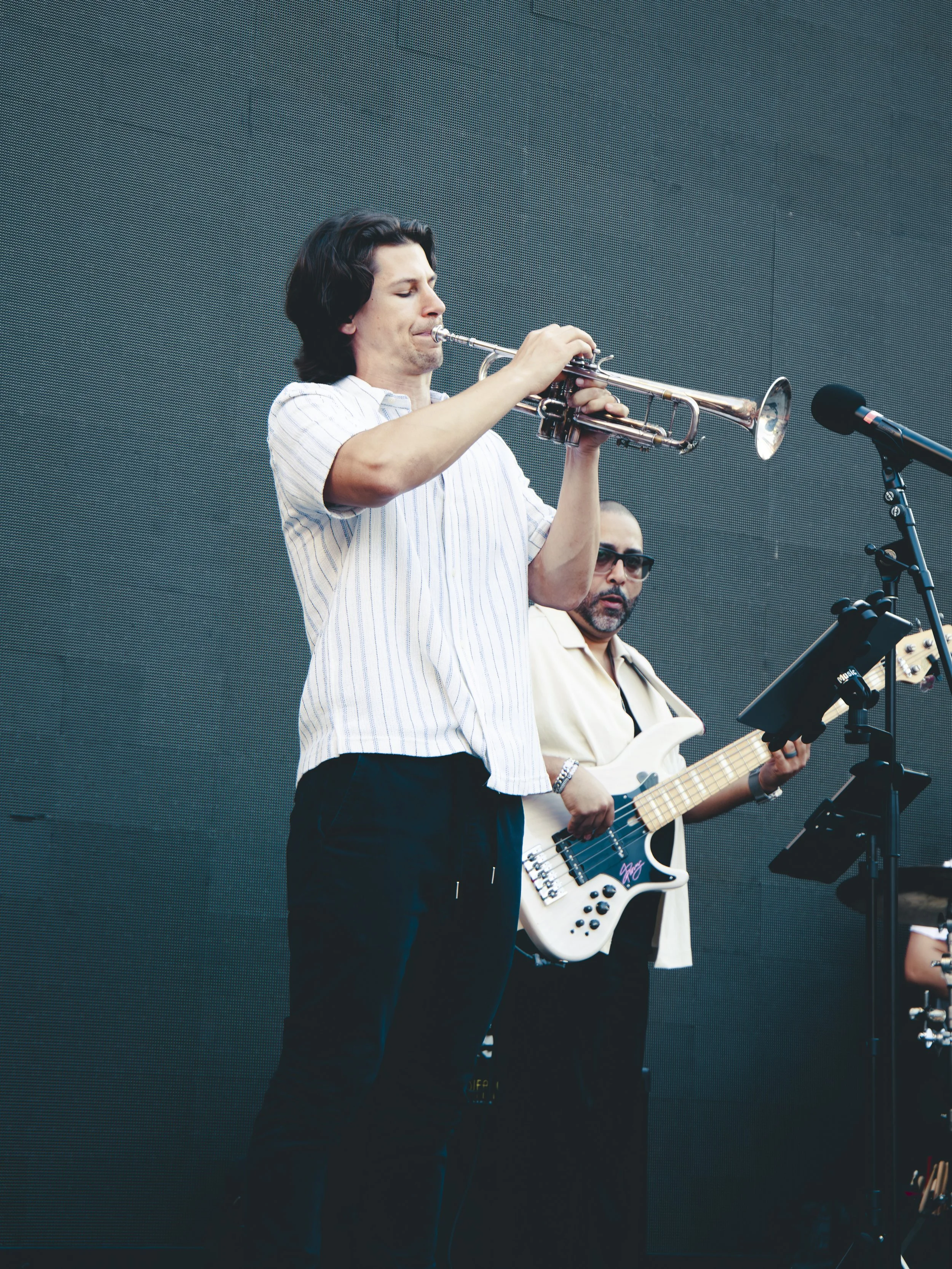 A man with dark hair playing a trumpet on stage, with another musician in the background playing a bass guitar.
