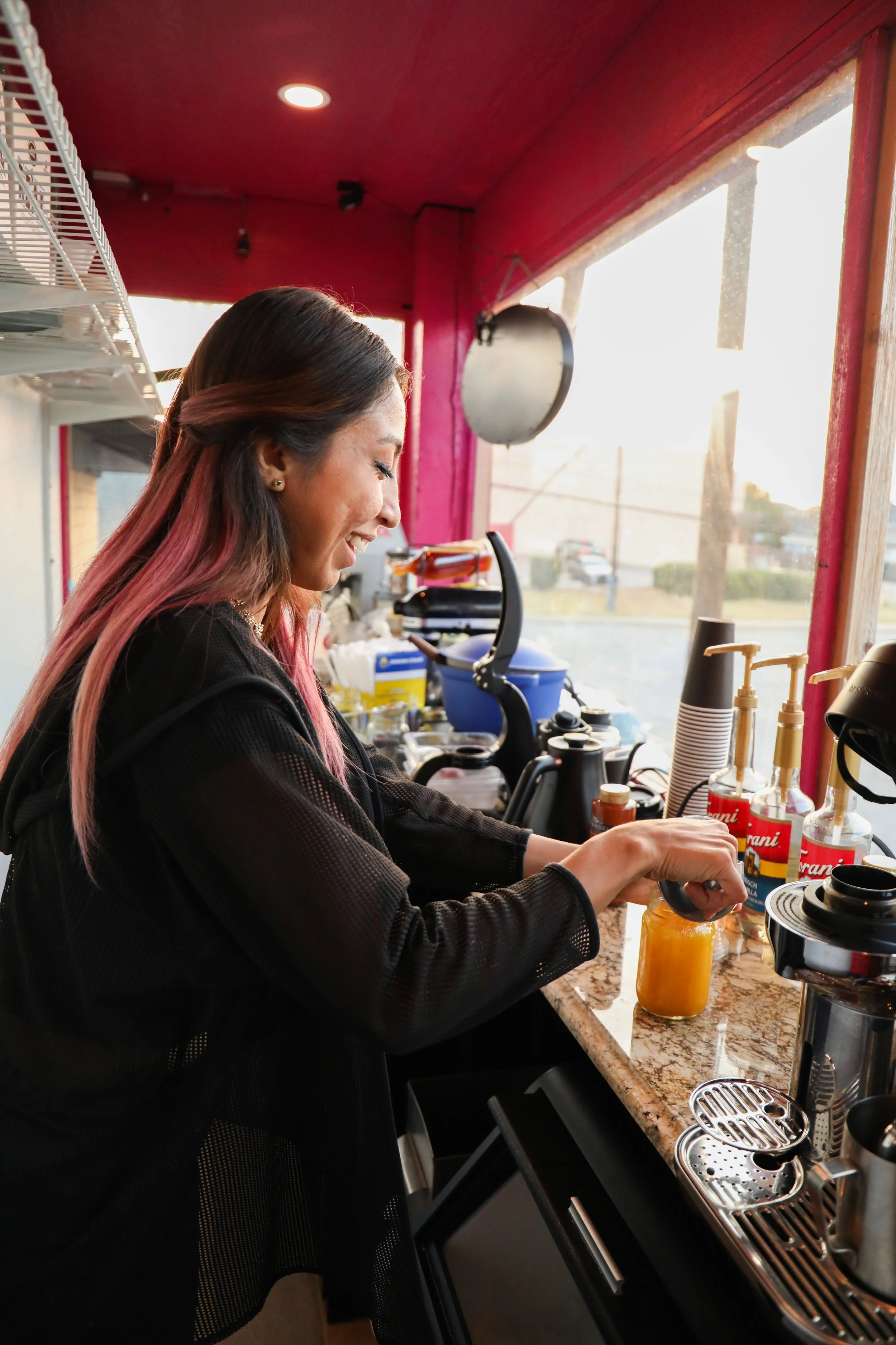 A woman with long hair dyed pink and brown, smiling while preparing a drink at a cafe counter, surrounded by bottles, equipment, and a coffee machine, with a window showing the outdoors.
