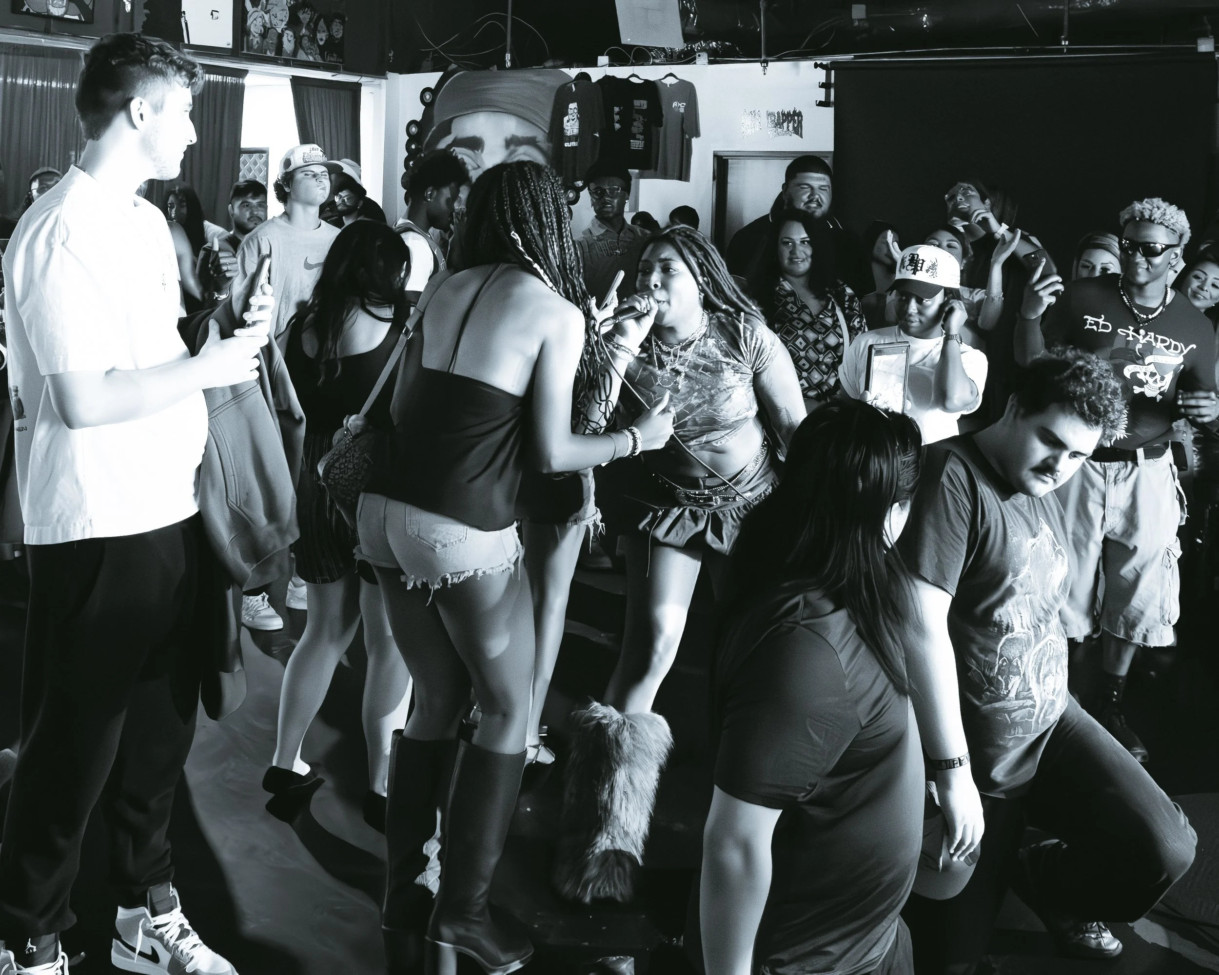 A diverse group of people gathered in a club or concert setting, some taking photos or videos, with a woman with braided hair singing into a microphone in the center.