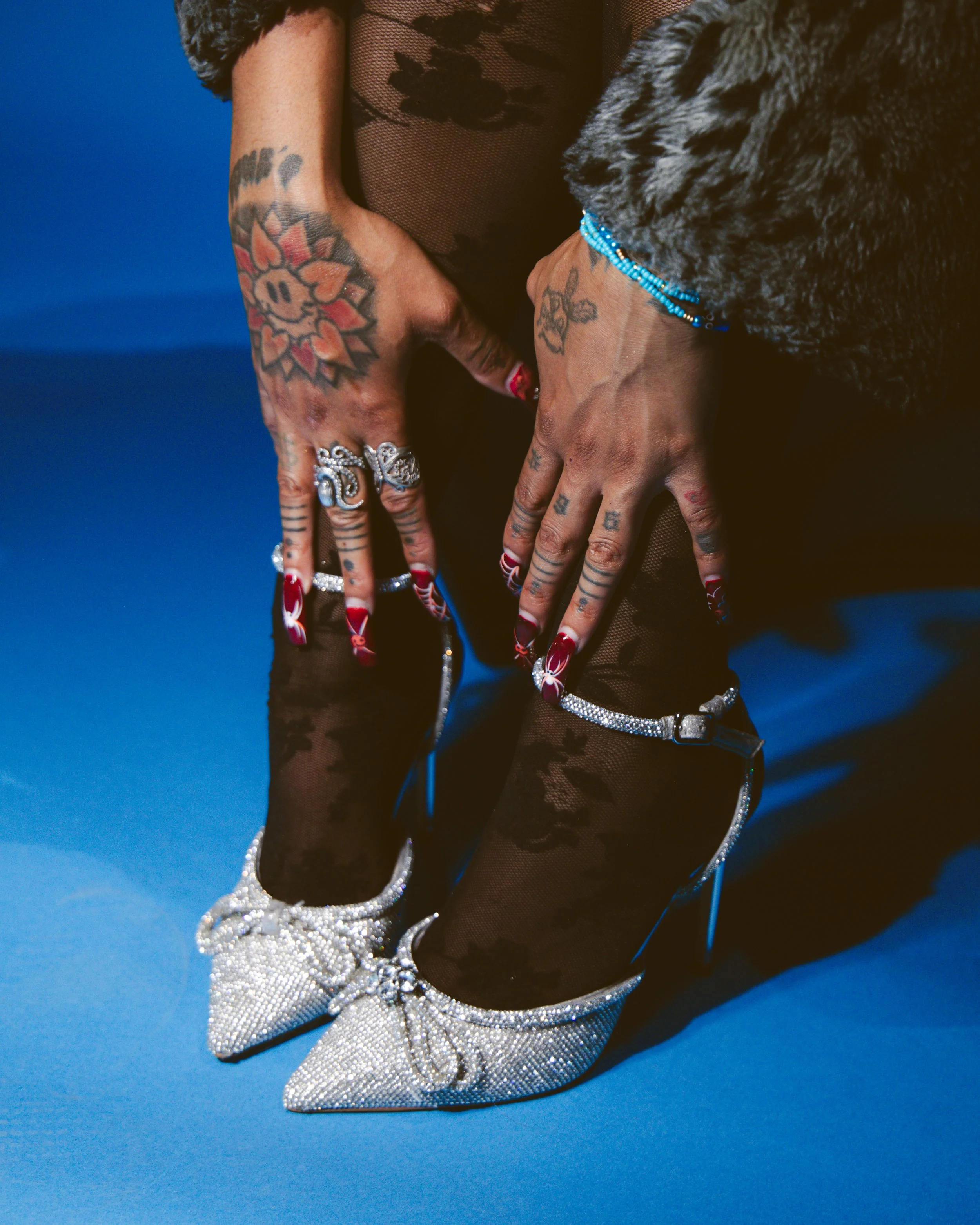 Close-up of a woman's hands with tattoos, wearing rings and red nails, resting on her lap. She is dressed in a black, lacy outfit with rhinestone trim, and black fishnet stockings. A large dog with curly fur and a blue beaded bracelet is partially vi
