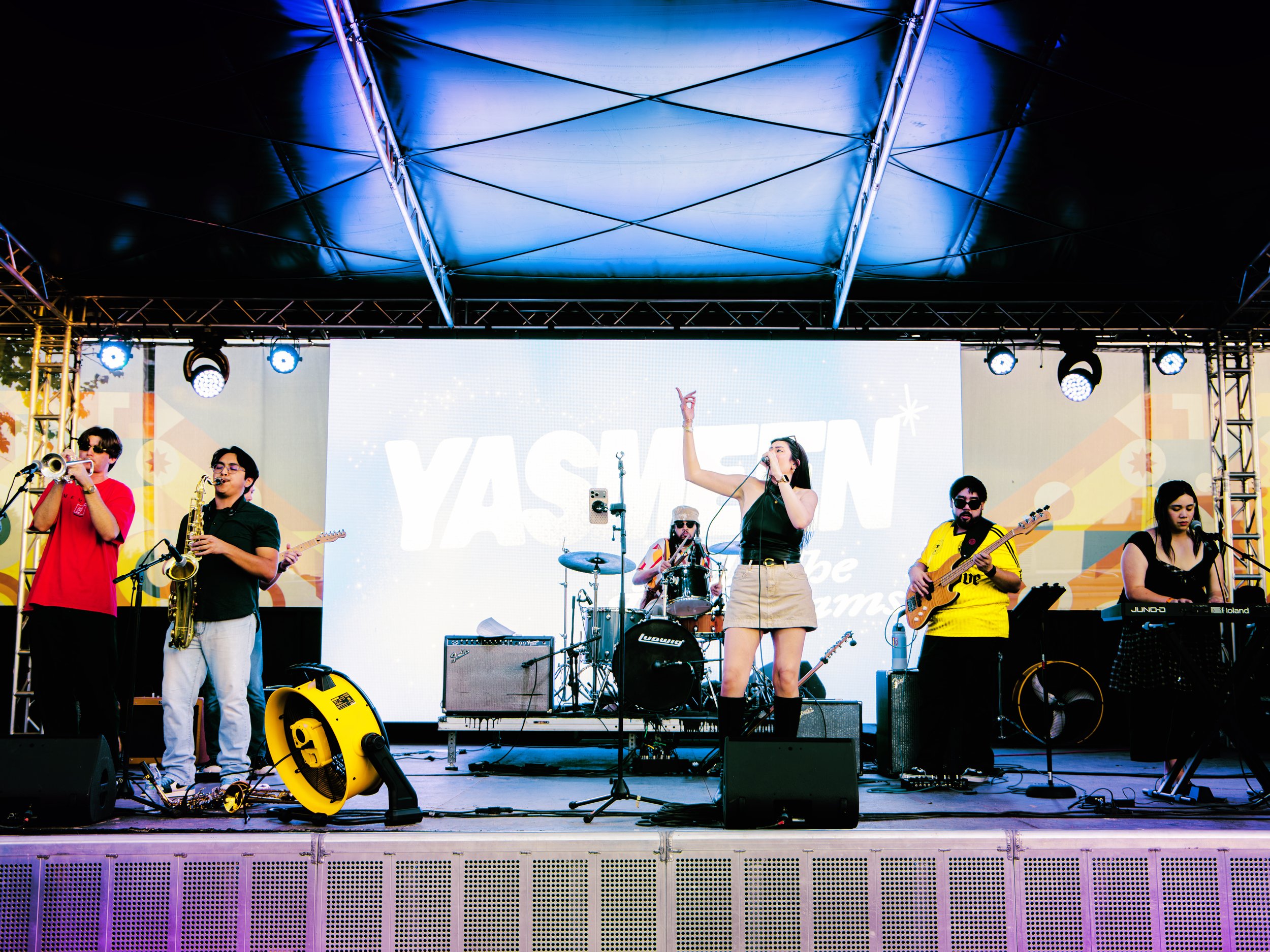 A group of musicians performing on a stage during an outdoor event at night, with a large LED screen behind them displaying the word "YASAN".