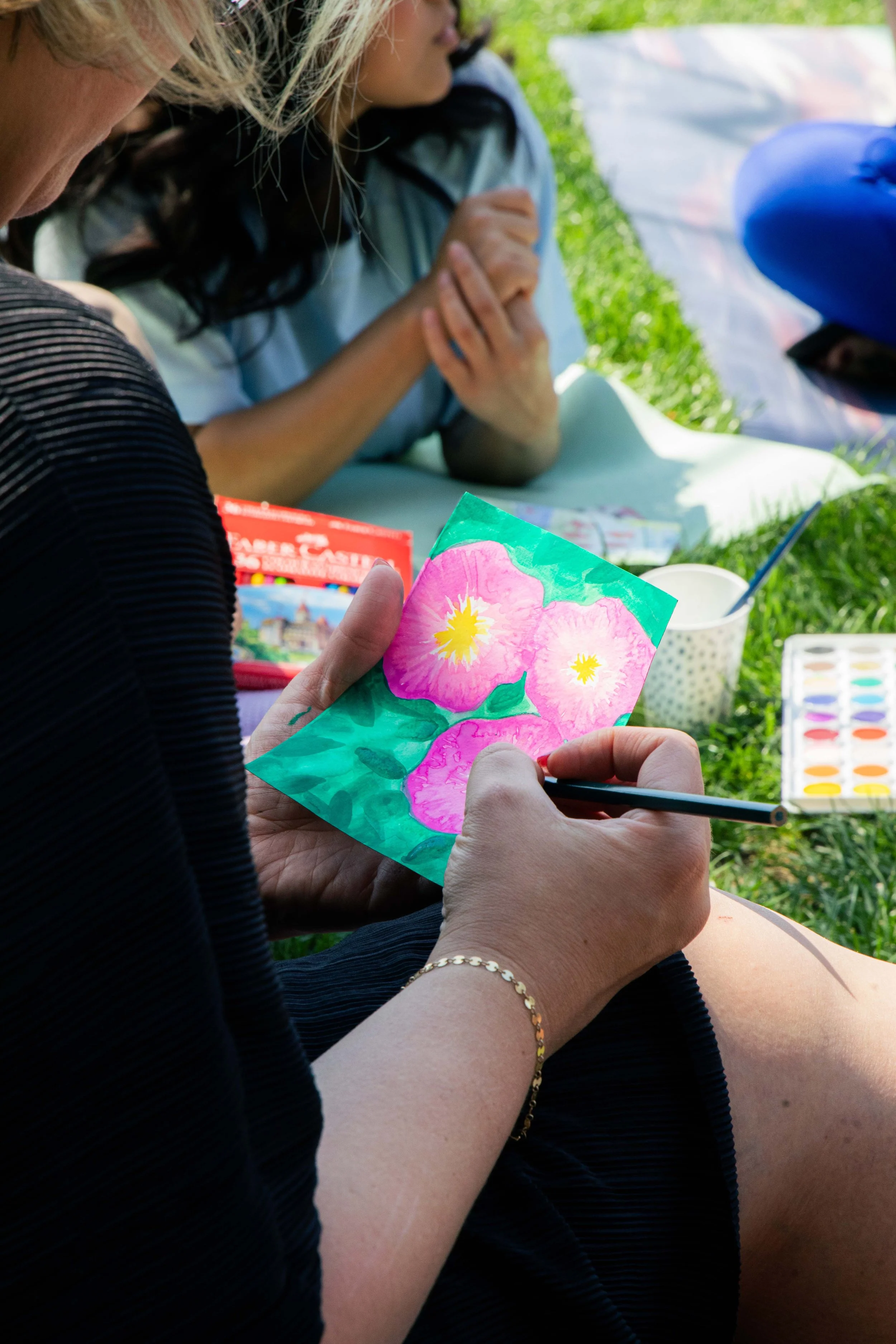 A person holding a colorful painted paper with pink flowers and green background, while other people are in the background on a grassy outdoor setting.