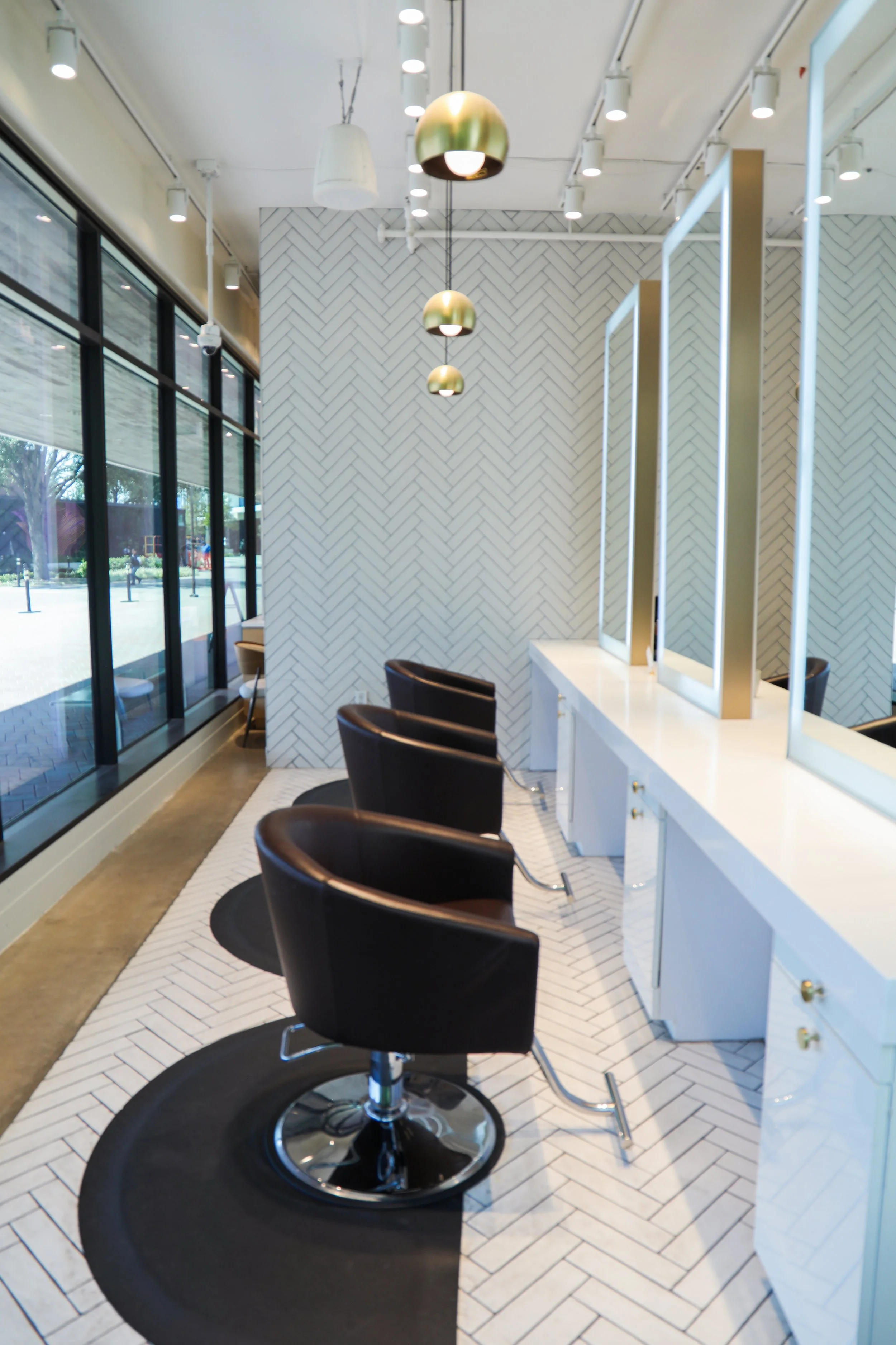 Empty hair salon with four styling chairs in front of mirrors, white counters, and a white herringbone tile wall. Large windows on the left let in natural light, illuminating the space.